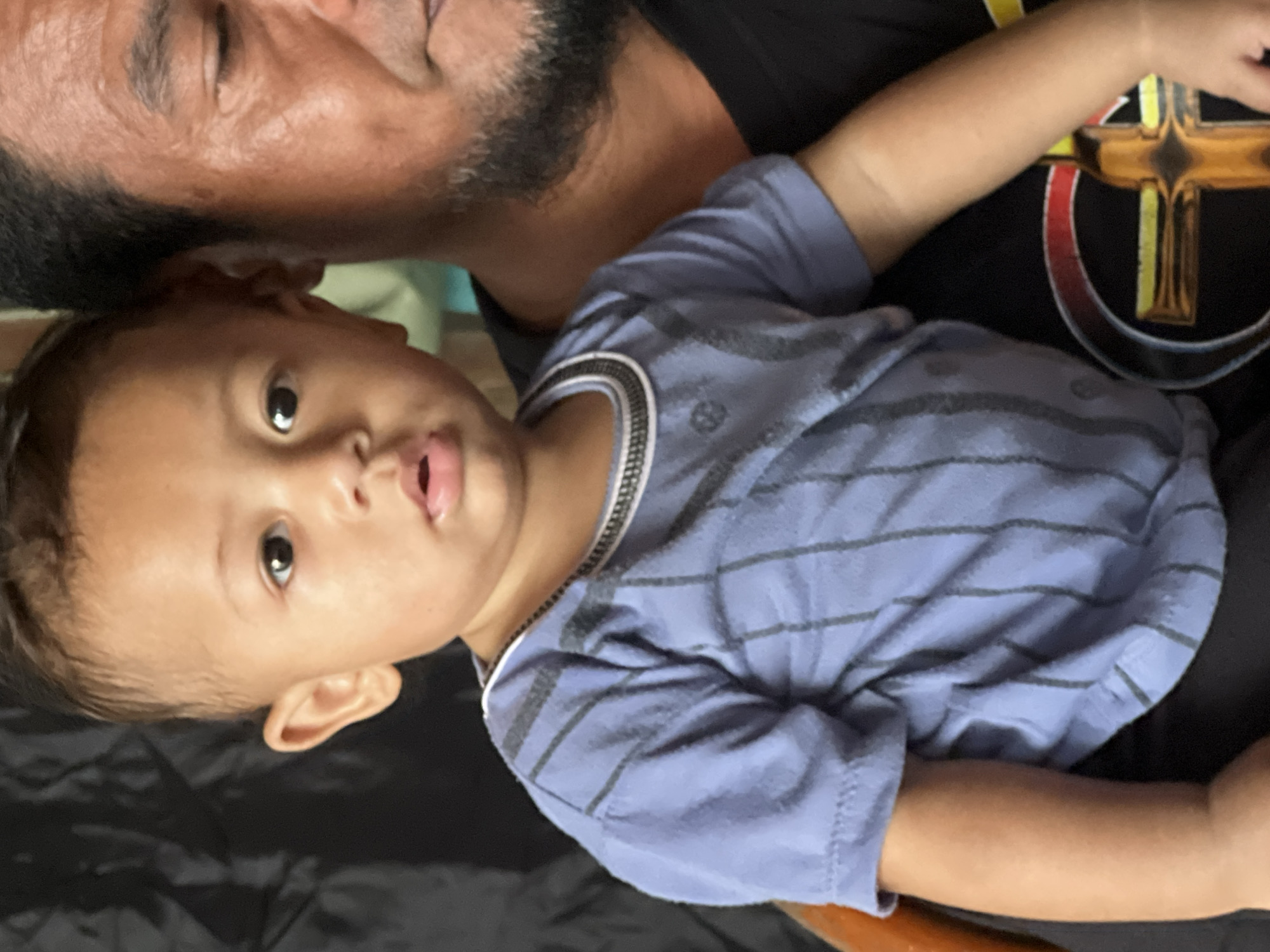 A close-up, medium shot captures a toddler held by an adult man. The toddler, facing forward, has dark hair and is wearing a blue t-shirt with dark gray vertical stripes and a white and gray striped neckline. The adult man, partially visible on the right, has dark hair and a beard, and is wearing a black t-shirt with a design featuring a cross. The background is dark and appears to be a textured fabric. The scene is set indoors, in Denpasar, Indonesia.