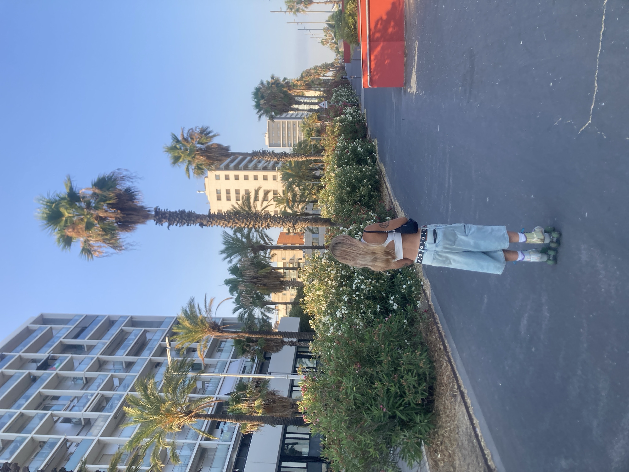 An outdoor, eye-level, full shot shows a young woman with blonde hair from behind, wearing a white crop top, light blue denim shorts, white socks, and roller skates. She is standing on a paved path lined with lush green bushes with white and pink flowers. Behind the bushes, there are tall palm trees and modern white buildings with balconies. The path continues into the distance, with more palm trees and buildings on the left side, and red structures on the right. The sky is clear and blue. The scene appears to be a promenade or boardwalk in Barcelona, Spain, likely in a coastal or park area, with architectural elements suggesting a Mediterranean urban environment.