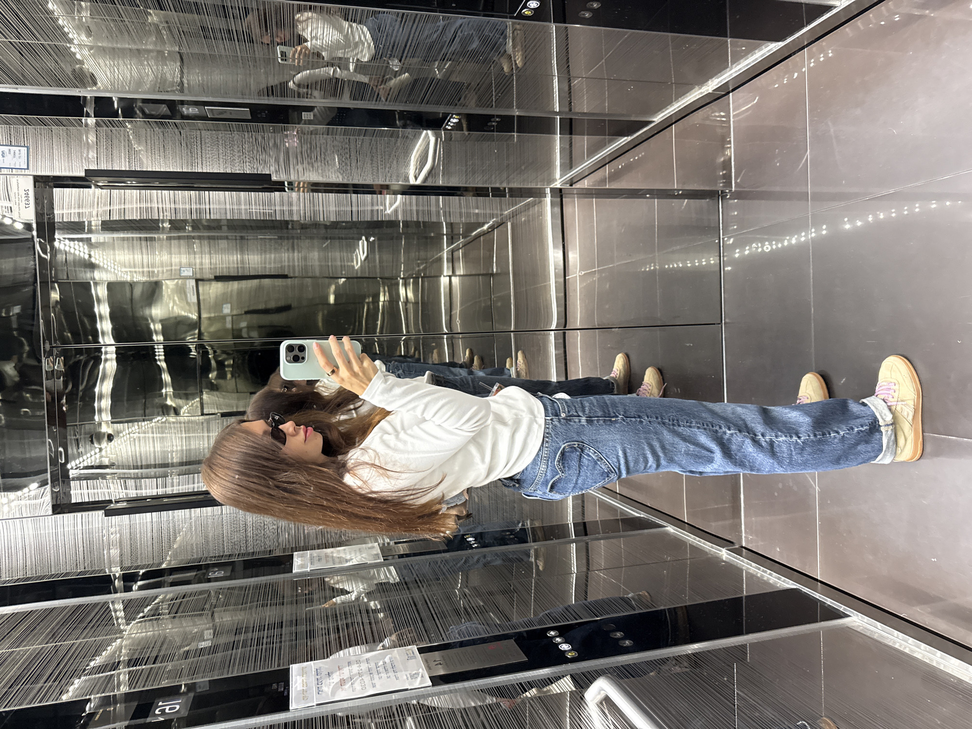 A woman is taking a selfie in an elevator located in Ramat Gan, Israel. The elevator has mirrored walls and a dark tiled floor. The woman has long brown hair, sunglasses, and is wearing a white top and blue jeans. She is holding a smartphone with a light green case to take the photo. Reflections of the woman and the elevator interior are visible in the mirrored walls.