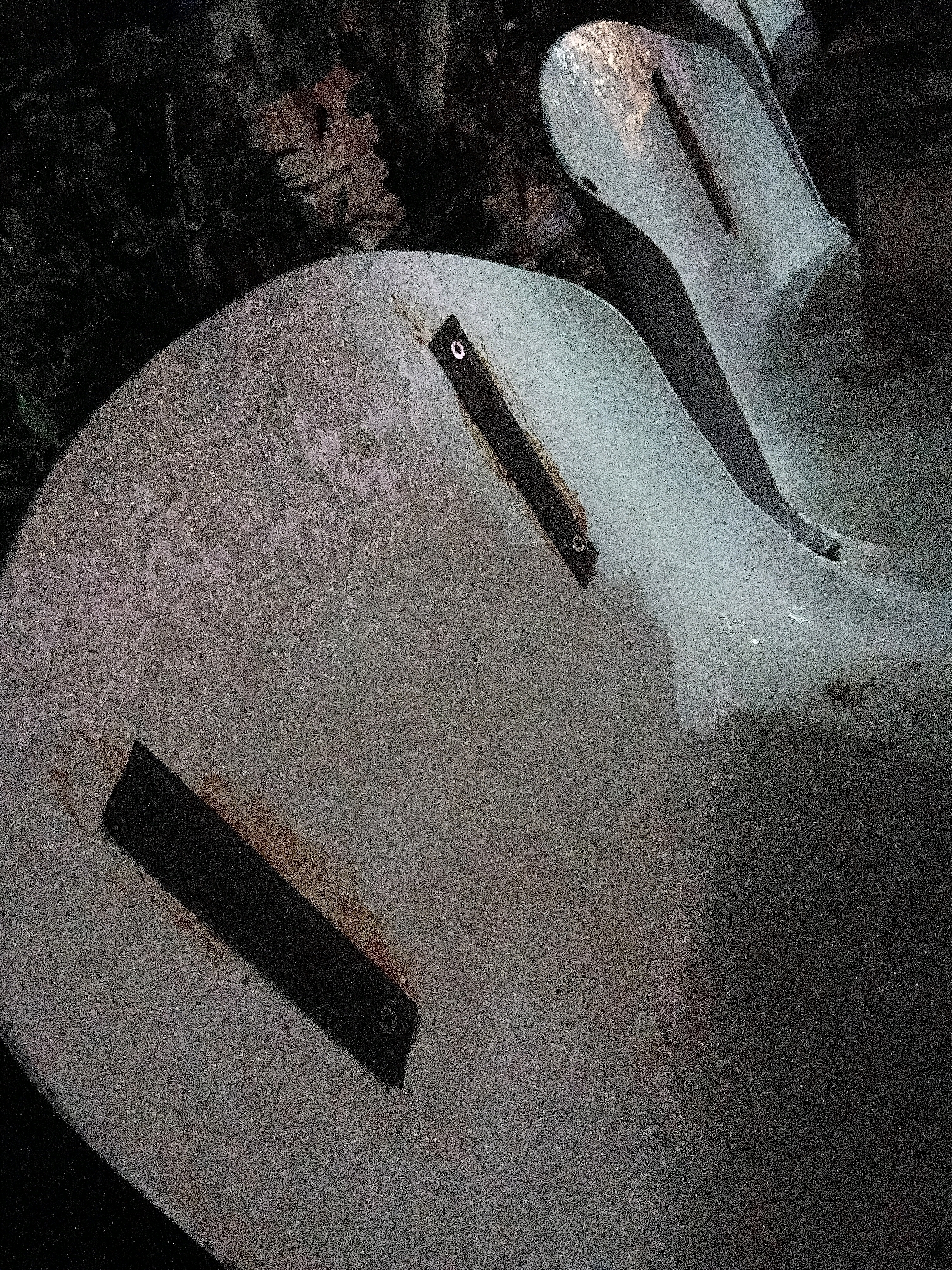 The image shows a section of a weathered, possibly concrete, slide at night. The slide appears to be a light grey color with a mottled texture. There are two dark, rectangular metal plates attached to the slide's surface, each with a visible circular fastener. The surrounding environment is dark and appears to be overgrown with foliage, suggesting an outdoor location. The lighting is dim, indicating it is likely nighttime. There are no people or animals visible in the image. The specific location in the city of Cikal, Indonesia, cannot be determined from the visual information provided.