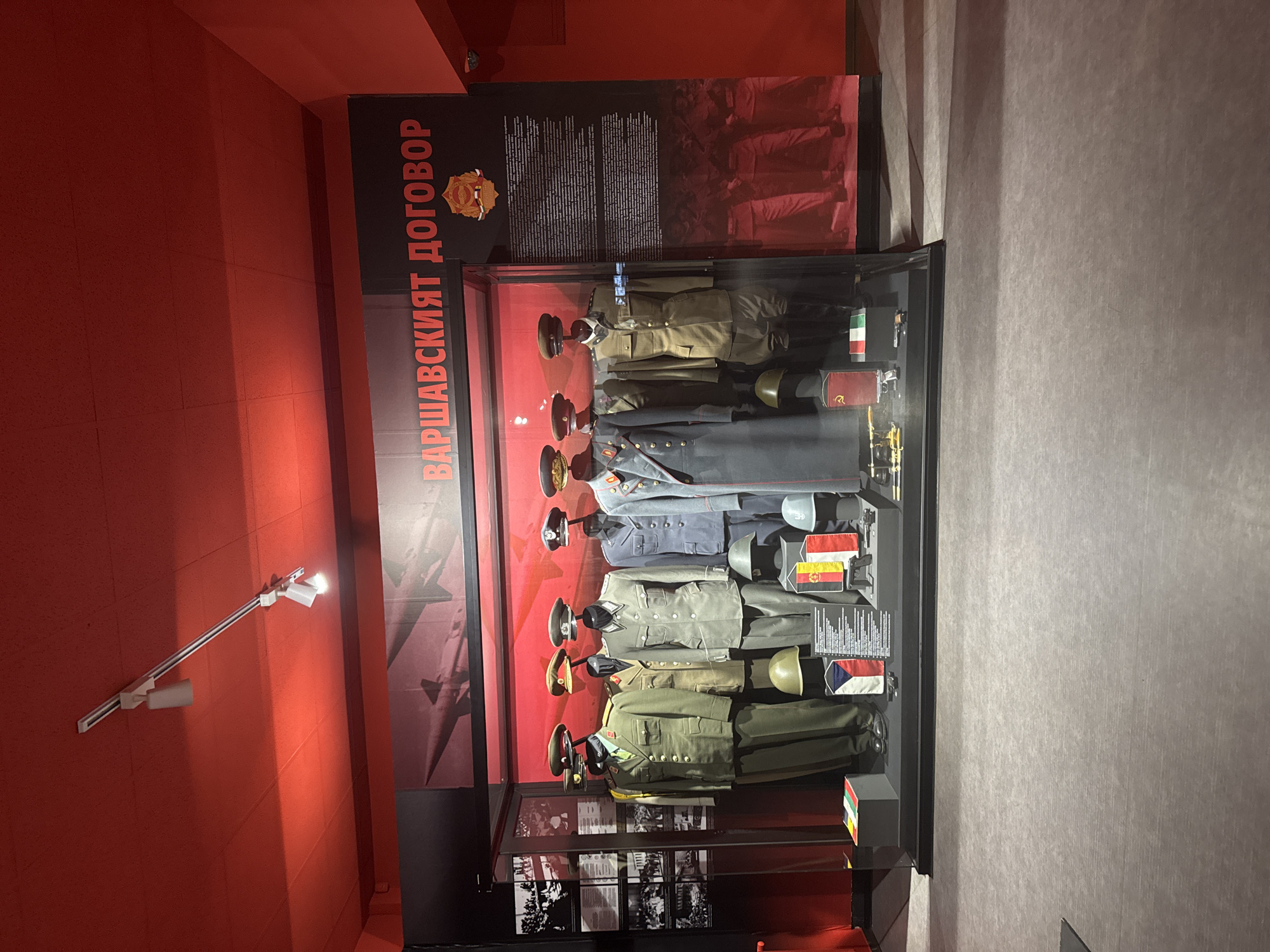 The image captures a display at the National Museum of Military History in Sofia, Bulgaria. The exhibit showcases military uniforms from Warsaw Pact countries, arranged in a glass display case. Behind the case, a large sign reads "ВАРШАВСКИЯТ ДОГОВОР" (Warsaw Pact) in Cyrillic script. The wall behind the display is painted red. The image is rotated.