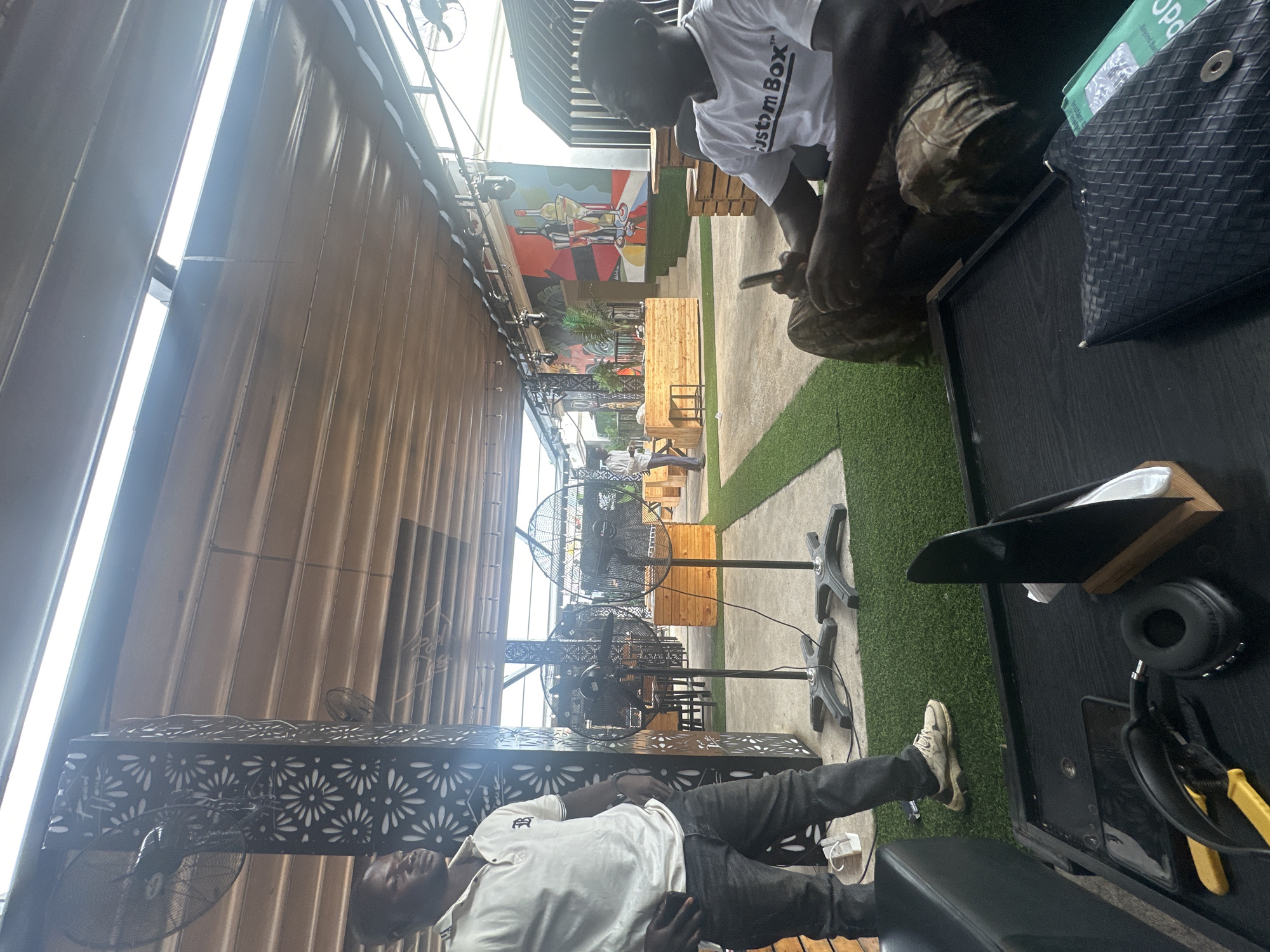 In Howayi, Nigeria, this image captures an outdoor seating area, possibly a restaurant or lounge, with a covered roof and artificial turf flooring. Two men are present. One man, wearing a white polo shirt and dark jeans, stands near a decorative metal pillar with a fan. The second man, in a white t-shirt with "Custom Box" printed on it and camouflage pants, is seated on a couch. Several large standing fans are positioned around the space, and wooden crates are used as seating or decor. A colorful mural depicting bottles and glasses adorns a distant wall. On a black table in the foreground, headphones, a phone, and other items are visible.