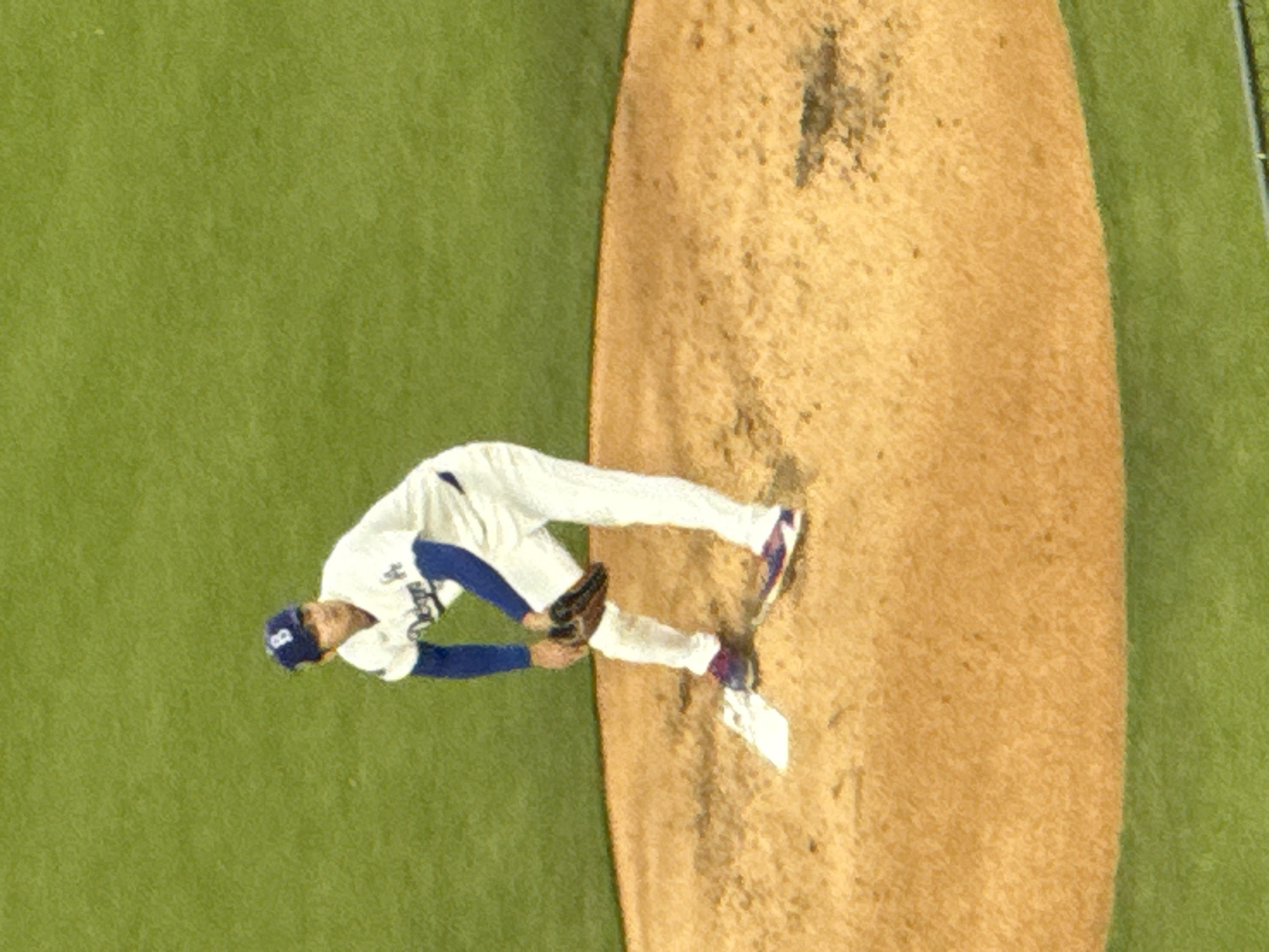 A baseball player in a white uniform with blue trim and the word "Dodgers" visible on the jersey, stands on the pitcher's mound during a game, likely at Dodger Stadium in Los Angeles. He wears a baseball cap with a "D" logo. He's in the middle of his pitching motion, with his glove extended and body angled towards the batter. The pitcher's mound is dirt. The background is bright green. A section of what appears to be a metal fence can be seen in the top right corner.