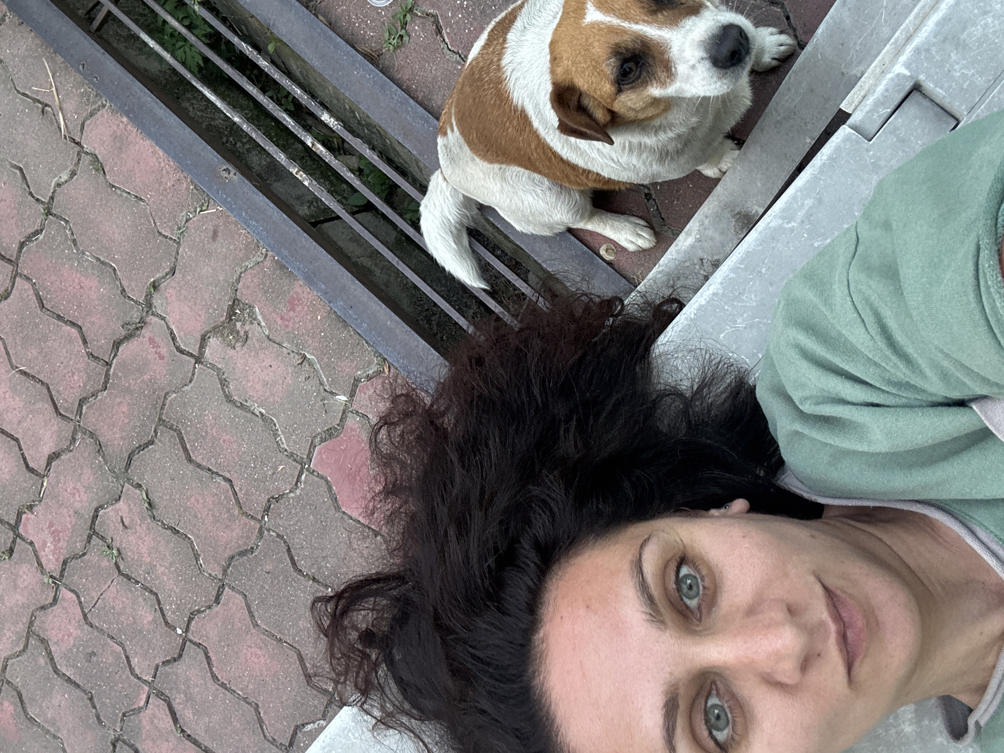 A close-up, high-angle selfie features a woman with dark, wavy hair lying down and looking at the camera. She is wearing a light green sweatshirt. Beside her, a Jack Russell Terrier with brown and white markings is sitting and looking towards the camera. They are situated on a red brick paver patio with a metal grate and railing to the right of the dog. The location is identified as Vesyoloye, Russia.