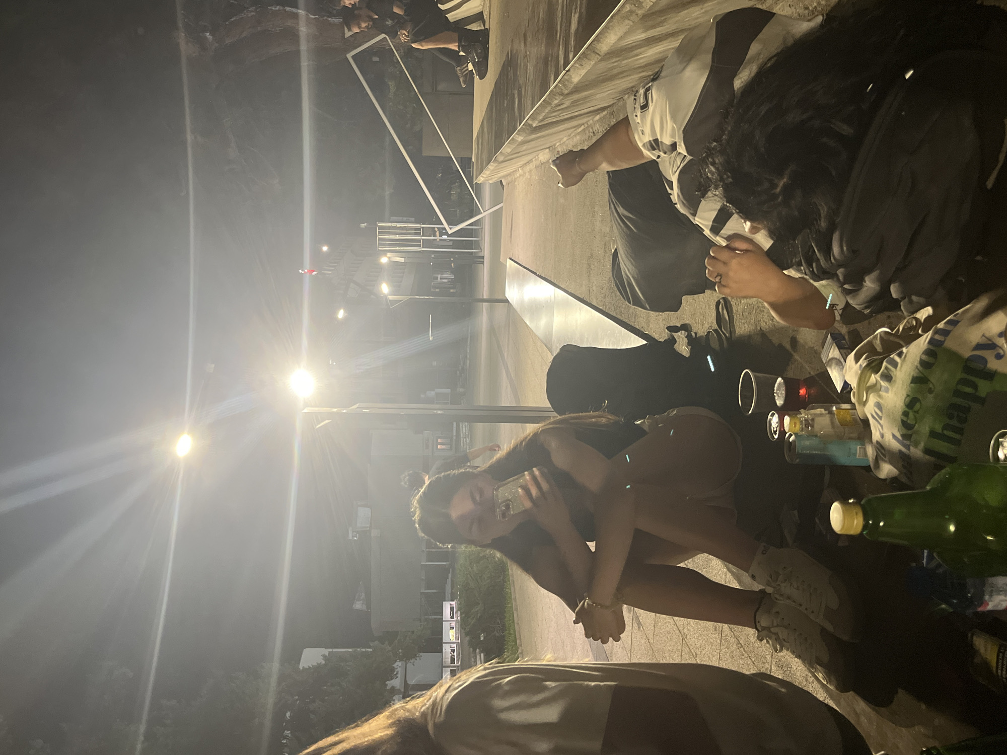 The image depicts an outdoor scene at night, possibly in a public area with streetlights illuminating the space. In the foreground, a young woman with long brown hair is sitting on the ground, holding a smartphone. She is wearing a dark top and light-colored shorts, with white sneakers on her feet. Her arms are crossed in front of her knees. To her left, another person, mostly out of frame, is also partially visible.
Scattered on the ground in front of the woman are several bottles and cups, some containing liquids of various colors, suggesting a social gathering. Behind the woman, a black backpack is placed on the ground. Further in the background, other individuals are visible, some sitting and others standing. One person is seen sitting on a raised platform with another person beside them. The environment appears to be an urban setting with buildings and streetlights visible in the distance. There are trees and foliage present, indicating a potentially park-like or landscaped area within the city.
The overall impression is a casual, nighttime gathering of young people. The forensic details would focus on the objects present and the arrangement of people, without making assumptions about their relationships or the exact nature of the event. The presence of smartphones, beverages, and the relaxed postures of the individuals suggest a social interaction.
