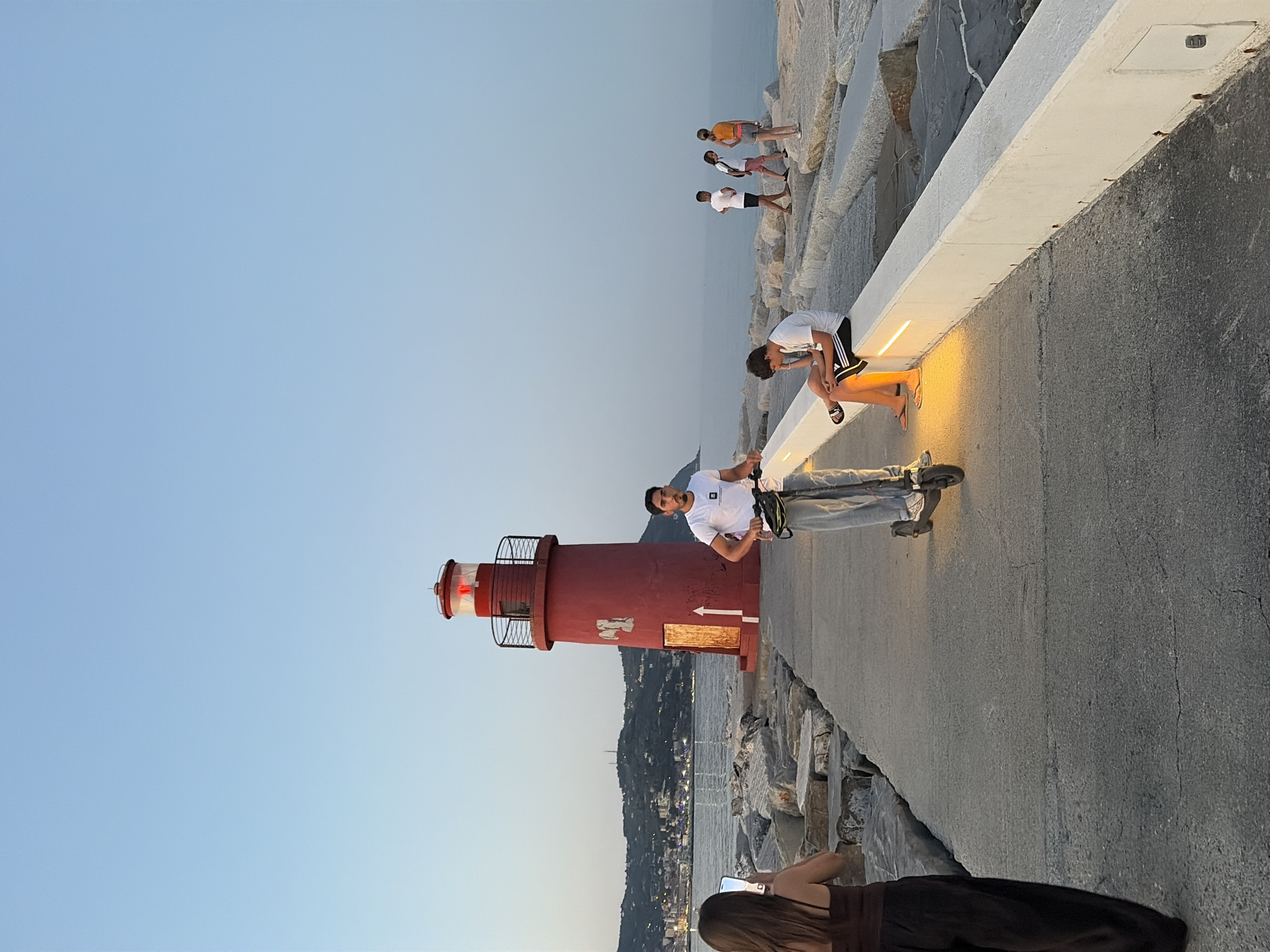 The image shows a group of people on a paved walkway next to a rocky breakwater. In the foreground, a red lighthouse with a black railing and a red light is visible. A man in a white t-shirt and jeans is standing next to the lighthouse, holding a selfie stick. A woman in a dark top is in the lower right corner of the image, taking a photo with her phone.
Further down the walkway, two young men are sitting, one in a white t-shirt and black shorts with Adidas stripes, and another in a white t-shirt and jeans, both illuminated by a warm yellow light. In the background, several other people are walking or standing on the breakwater. The scene is set against a clear blue sky, suggesting it is daytime, likely late afternoon or early evening given the warm lighting. The presence of the lighthouse, breakwater, and the distant town with lights suggests a coastal location. The water is calm. The overall mood appears relaxed and casual, with people enjoying the scenery and taking photos. The location context of Imperia, Italy, further supports the coastal setting.