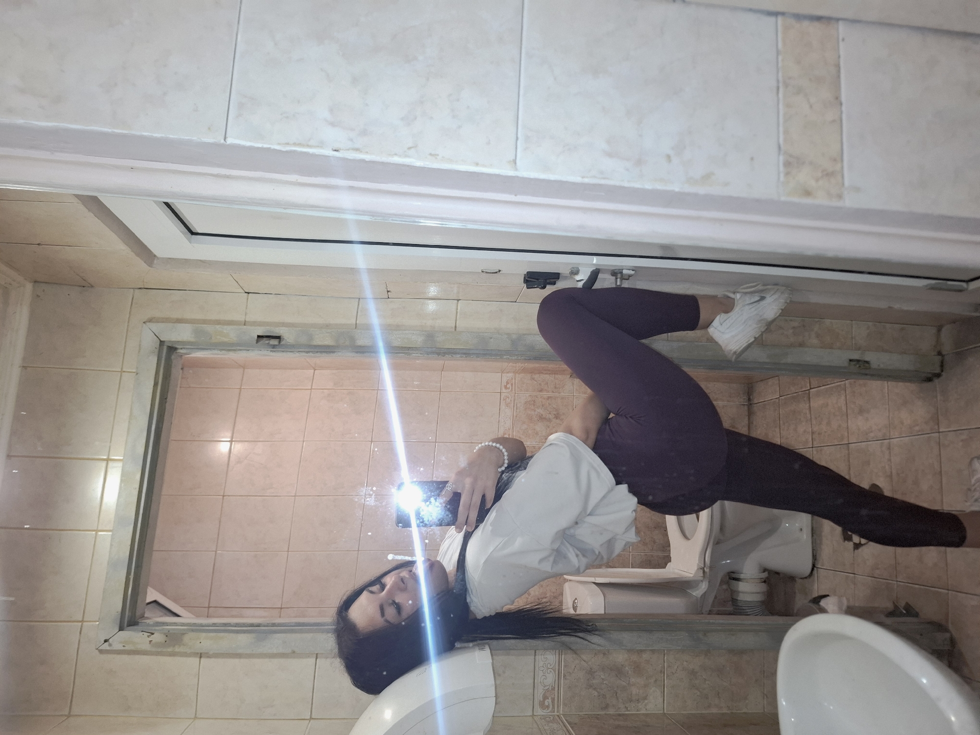 A young adult female with long dark hair is depicted in a mirror selfie taken within a tiled restroom located in Mezdra, Bulgaria. She wears a white cropped t-shirt, dark purple full-length leggings, and white athletic shoes with a visible Nike swoosh on the right foot. Her right hand holds a black smartphone, from which a bright flash emanates, creating a strong horizontal light streak across the image. A pearl bracelet is on her right wrist, and a ring is visible on a finger of her right hand. She is posed with her left leg bent, resting her white-clad foot against a white doorframe on the right side of the image. Her torso is tilted towards the left. The restroom features light beige square wall and floor tiles, with a darker decorative tiled border. Visible fixtures include a white wall-mounted automatic hand dryer, a white ceramic sink (partially visible lower left), and a white ceramic toilet with an open lid in the background. A secondary doorway with a metal frame is visible behind the subject, leading to an interior area with similar, slightly pinker tiles. A black door handle mechanism is present on the right doorframe.