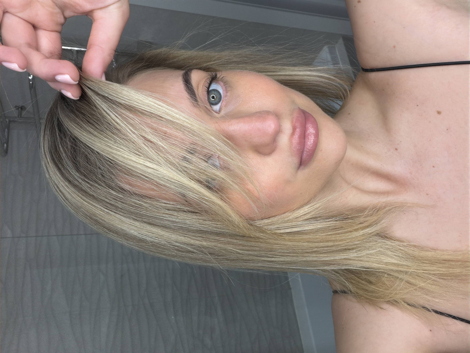 A close-up, rotated portrait shot depicts a young woman with blonde hair, looking directly at the camera. Her head is tilted, and her left hand is visible in the upper part of the frame, gently pulling a strand of her hair near the scalp. Her nails are manicured with light pink polish.
Her left eye, a striking light blue or green, is clearly visible, framed by dark, defined eyebrows. Her skin appears fair, with a subtle blush on her cheeks, and her lips are parted slightly, showing a soft pinkish-nude color. Her straight, blonde hair, with noticeable darker roots or lowlights, cascades across her face and over her right shoulder.
The woman's bare shoulder and upper chest are visible, revealing a few scattered freckles or moles. A thin black strap, likely from a top or camisole, crosses her shoulder. The background is a light gray wall with a subtle, repeating wavy or textured pattern, suggesting an indoor setting such as a bathroom. The overall image gives the impression of a posed photograph or selfie taken in Sofiivska Borshchahivka, Ukraine. There is no visible text in the image.