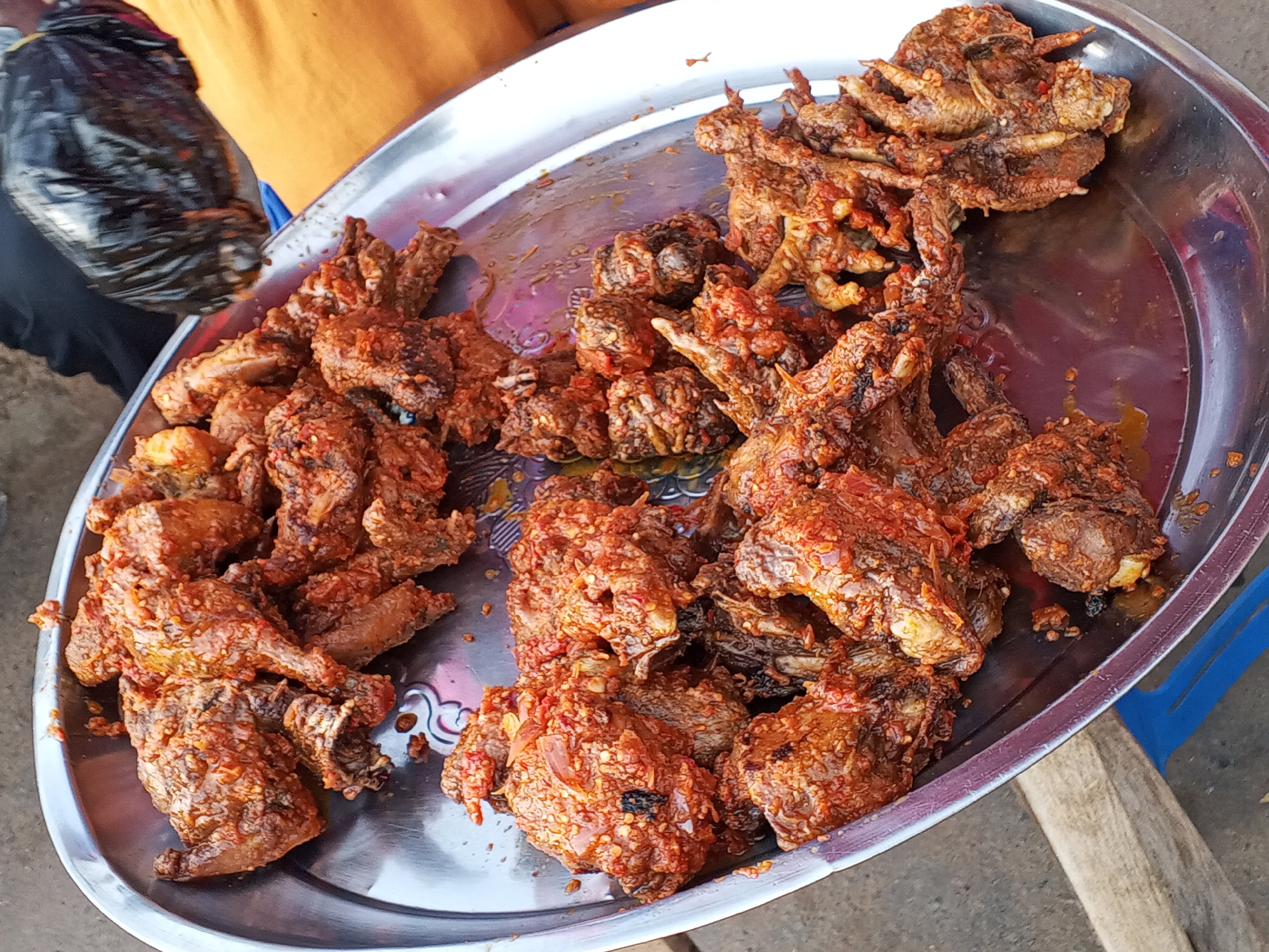 A close-up, eye-level shot shows a silver tray filled with spicy fried chicken pieces in Gwong, Nigeria. The chicken is covered in a reddish-brown sauce and is piled high on the tray. A person wearing an orange shirt and holding a black plastic bag is partially visible on the left side of the tray. Part of a blue plastic stool is visible on the right side of the tray. The background is slightly blurred.