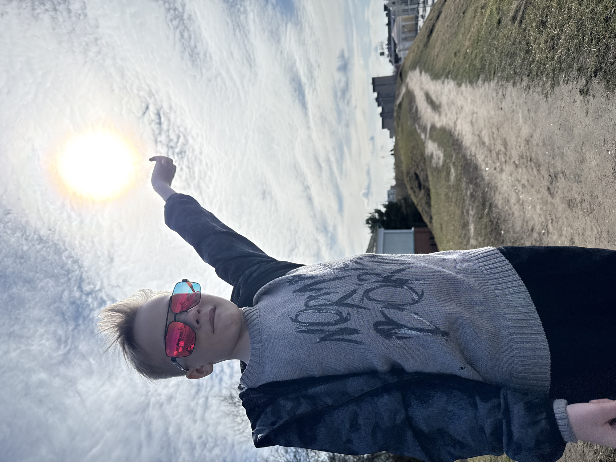 A young, light-skinned person is photographed outdoors in an off-kilter portrait. The person wears red-tinted sunglasses, a grey sweater, and a black jacket. They are reaching upwards towards the bright sun in the sky. The background includes cloudy skies, a dirt path, a grassy hillside, and buildings. The city is Dmitrov, Russia.