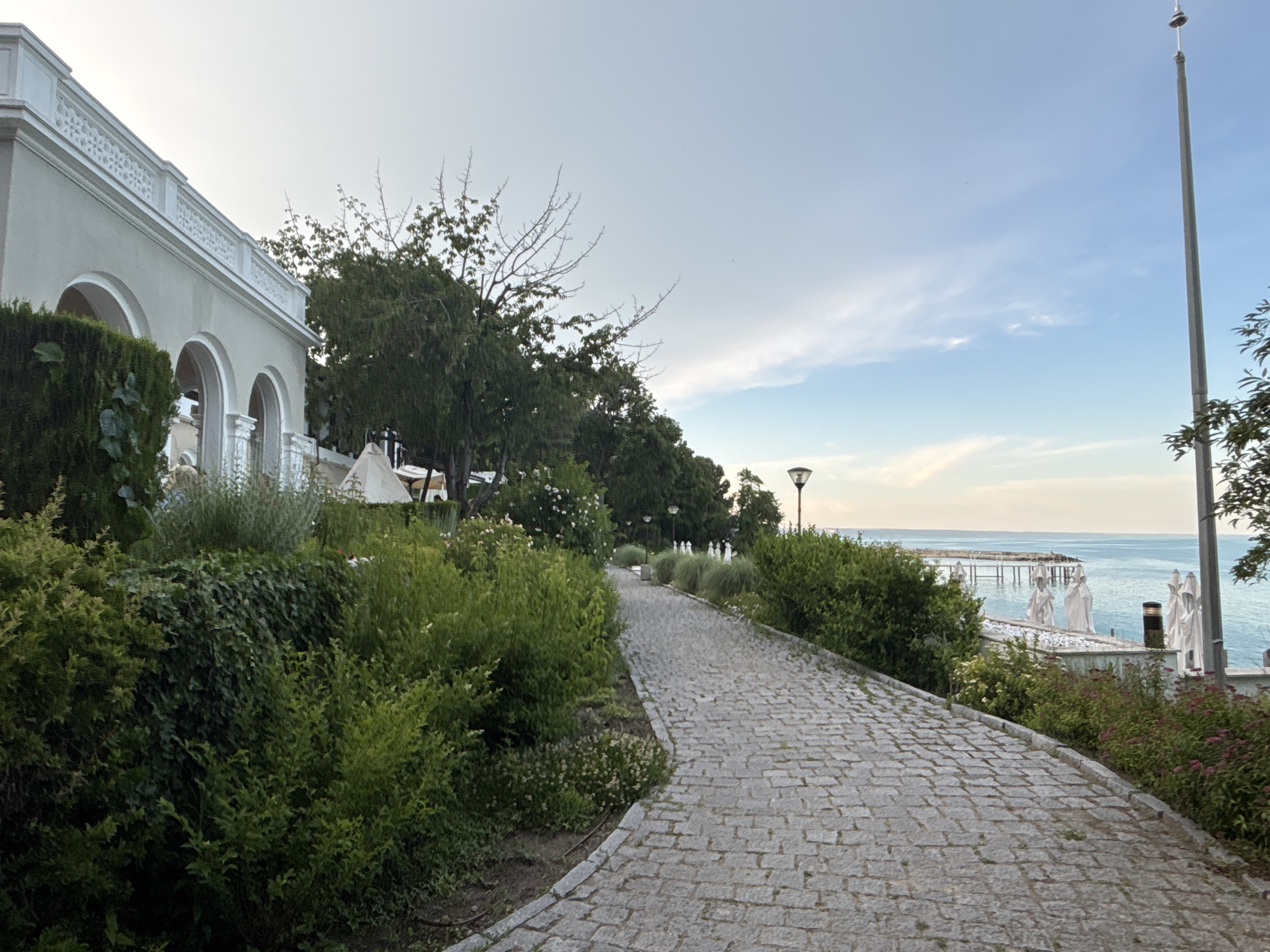 The image depicts a paved walkway lined with lush greenery, leading towards the sea. On the left, a light-colored building with arched openings and decorative railings is visible, partially obscured by hedges. The walkway is made of cobblestones and curves to the right, offering a view of the sea and a pier in the distance. Several white umbrellas are set up near the water. A tall flagpole stands on the right side of the image. The sky is partly cloudy with a mix of blue and white. This scene is located at the Sea Garden in Varna, Bulgaria, a prominent park overlooking the Black Sea. There are no people visible in the image.