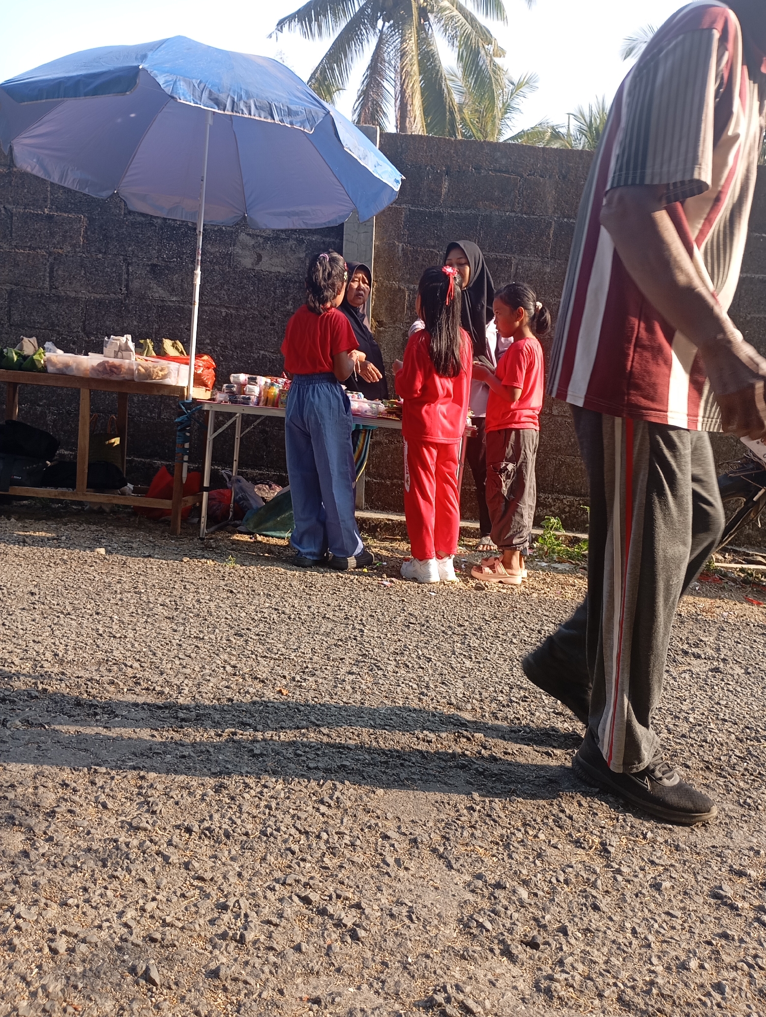 In Ketepung, Indonesia, an outdoor scene unfolds at midday with a stall set up under a large blue umbrella. The stall displays various food items in plastic containers and bags. Three young girls and two adult women are gathered near the stall, interacting with each other. One girl is wearing a red shirt and blue pants, another a red shirt and dark pants, and the third a red shirt. An adult woman in a black hijab is also present, along with another woman. In the foreground, a person is partially visible from the side, wearing a striped t-shirt and dark pants with red and white stripes. The ground is covered with gravel, and shadows are cast on it, indicating bright sunlight. A brick wall forms the backdrop, with palm trees visible against a clear sky.