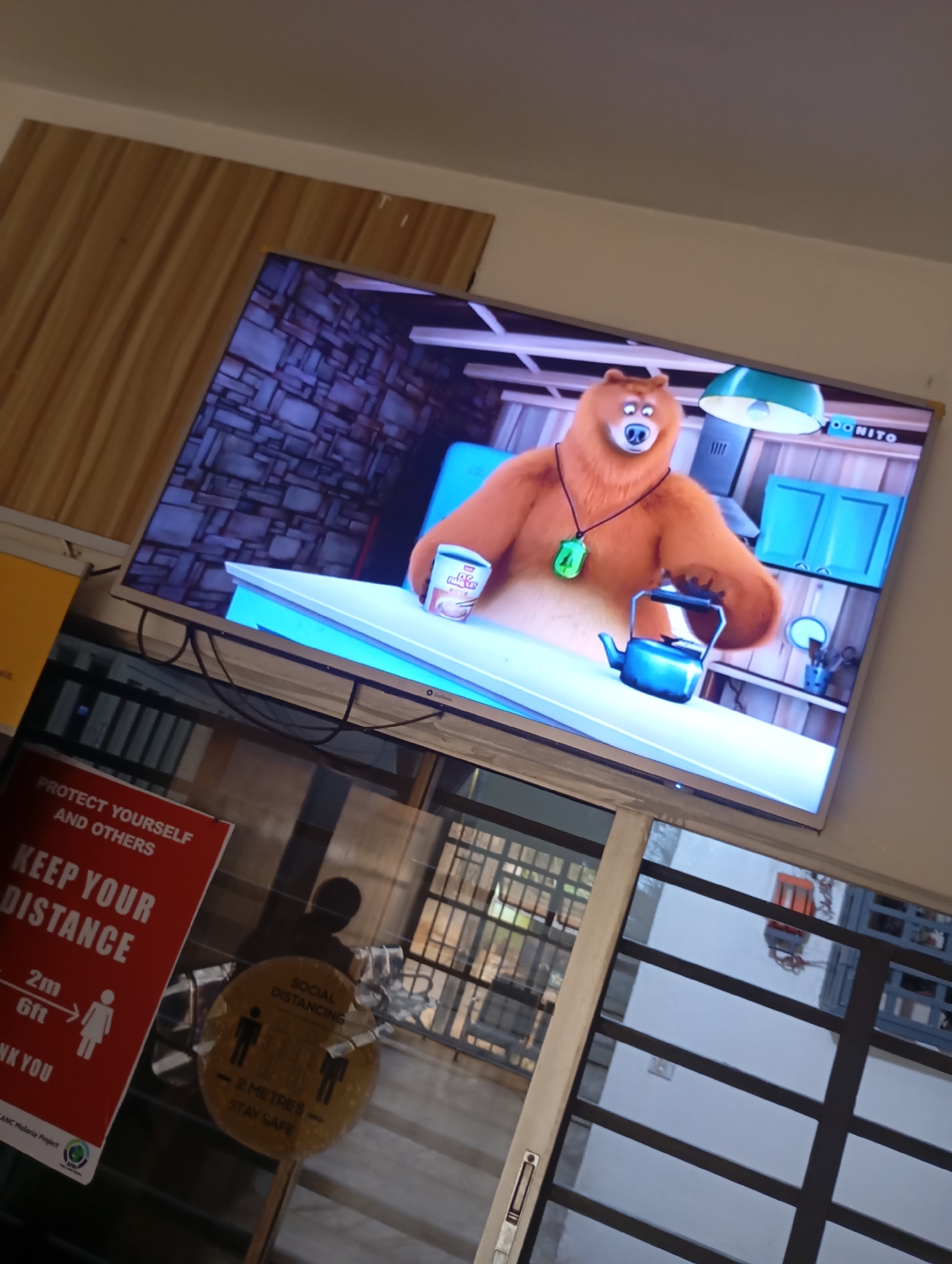 A television screen displays a cartoon of a brown bear wearing a green necklace with the number 4 on it, holding a can and a blue teapot. The bear is seated at a white desk. The TV is placed in a room with wood-paneled walls, a white ceiling, and a light-colored floor. In front of the TV, on the floor, is a red sign with white text that reads "PROTECT YOURSELF AND OTHERS KEEP YOUR DISTANCE 2m 6ft THANK YOU." Next to it, on the floor, is a circular yellow sign with black text and graphics illustrating social distancing. This scene appears to be in a public area, possibly a shop or office, in Jos, Nigeria, suggested by the presence of the safety signs. The lighting is artificial, with the television screen being the primary light source.