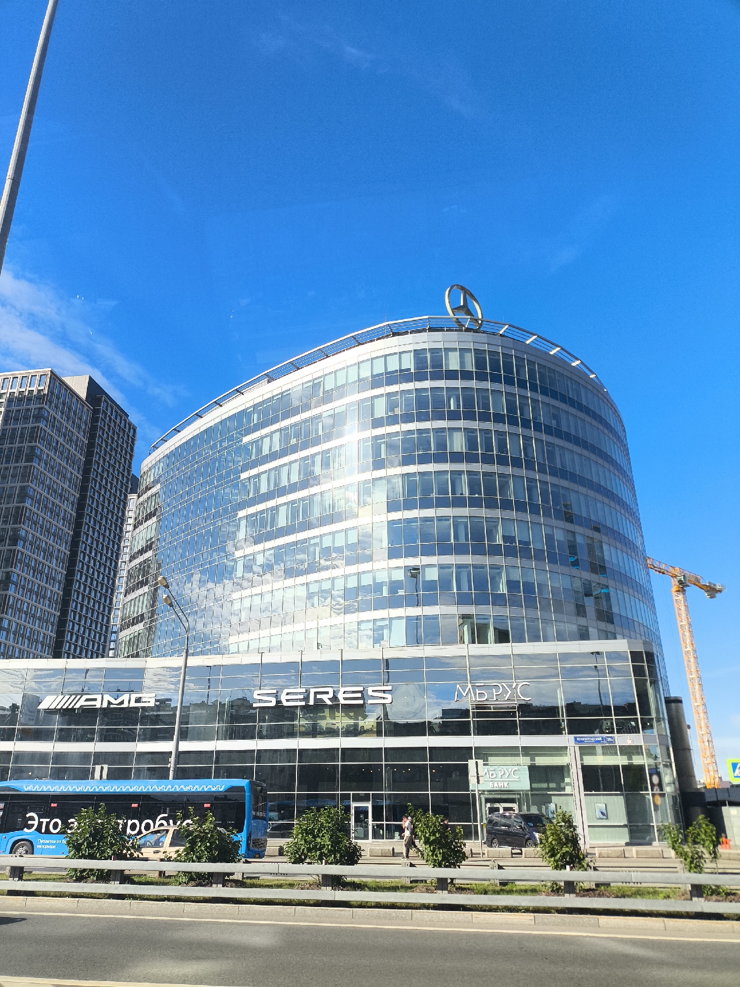 A low-angle, full shot captures a modern, curved glass building against a bright blue sky. A prominent Mercedes-Benz logo adorns the top of the building. To the left, a taller, rectangular building stands, also covered in windows. The facade of the curved building features large glass panels reflecting the sky and surrounding environment. Several white and silver signs are visible on the lower level, including "AMG," "SERES," and "МБРУС" (MB RUS) with "БАНК" (BANK) below it. A blue bus with white decorative elements and Russian text is parked in front of the building. Green bushes are planted along a roadside barrier in the foreground. A construction crane is visible in the background to the right. The scene appears to be in an urban environment.