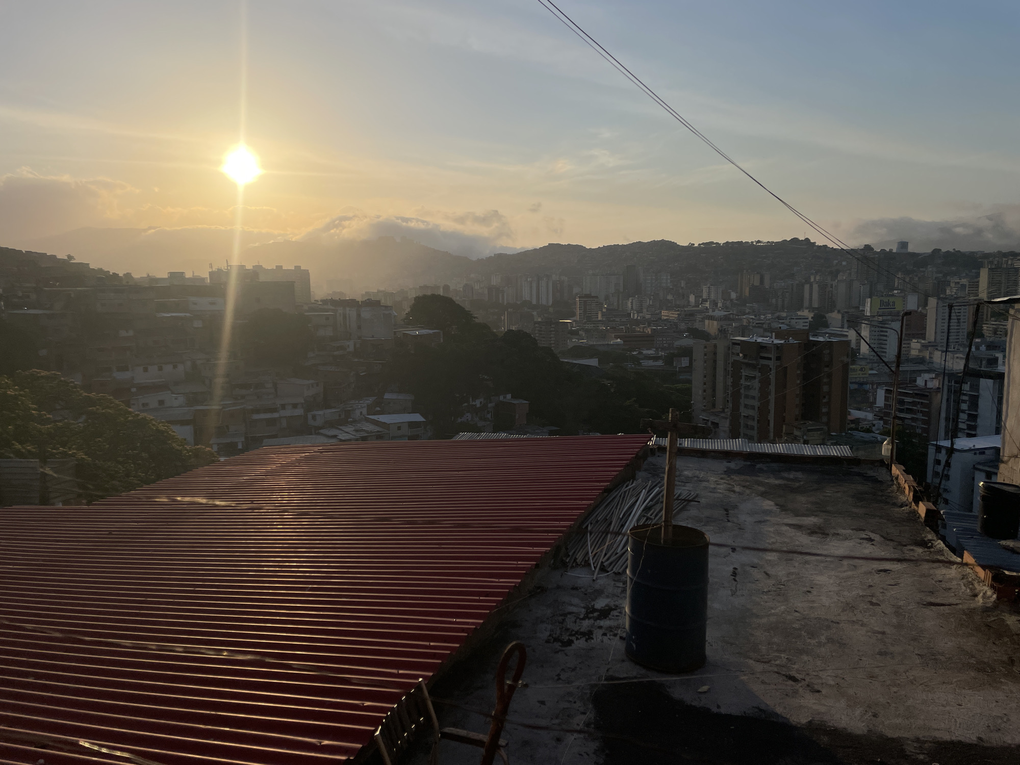 The image presents an elevated, wide-angle view of the city of Caracas, Venezuela, at either sunrise or sunset, with strong, direct sunlight emanating from the upper left quadrant. The immediate foreground on the right features a weathered, stained concrete rooftop. On this surface, a dark blue 55-gallon metal drum stands upright, containing several vertical wooden poles. Adjacent to the drum, a pile of white PVC pipes is stacked. A clean, corrugated red metal roof slopes from the right foreground towards the lower left. A reddish-brown metal structure, possibly part of a ladder, is partially visible in the bottom left. Two thin, dark cables or wires diagonally traverse the upper half of the sky.
The midground reveals a dense urban landscape, characteristic of Caracas, with a mix of multi-story buildings, including formal structures and informal settlements (barrios) sprawling across numerous hills and into valleys. Patches of trees and vegetation are interspersed among the buildings. A large billboard with the text "Daka" is visible on a building in the mid-right. The background is dominated by mountain ranges partially shrouded in atmospheric haze and clouds, particularly near the sun's position. The sky transitions from a bright yellow-orange near the sun to a pale blue at the top right. No people or specific actions are discernible within the frame.