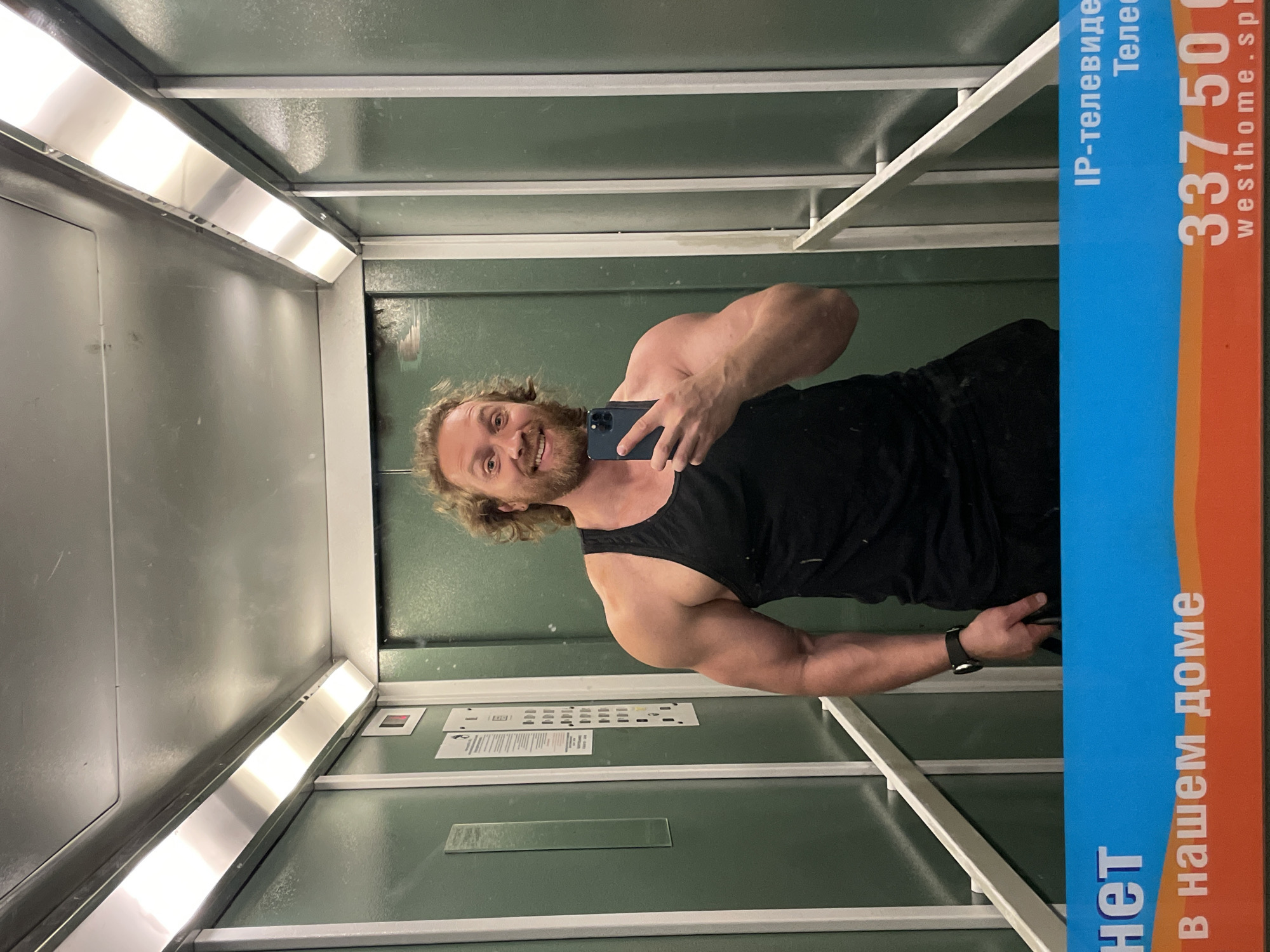 This is a vertical, slightly blurry selfie taken in an elevator. The main subject is a man with long, wavy blonde hair and a beard, smiling at the camera while holding a dark-colored smartphone. He is wearing a black tank top and a black wristwatch on his left arm. His muscular arms and shoulders are visible.
The elevator interior is utilitarian, with green walls and silver metal trim. Fluorescent lighting fixtures are visible along the top. There is a poster or advertisement on the right side of the elevator, vertically oriented, with text in both English and Russian. The visible text includes "IP-телевидение" (IP television), "Телес" (Tele...), "337 500", and "westhome.spl". Below this, in larger text, it reads "в нашем доме" (in our home) and "нет" (no/not).
The man appears relaxed and happy. The overall impression is a casual moment captured in an everyday setting. The scene is indoors, likely in a building in Saint Petersburg, Russia, as indicated by the Russian text on the advertisement. The time of day and weather are not discernible from the image.