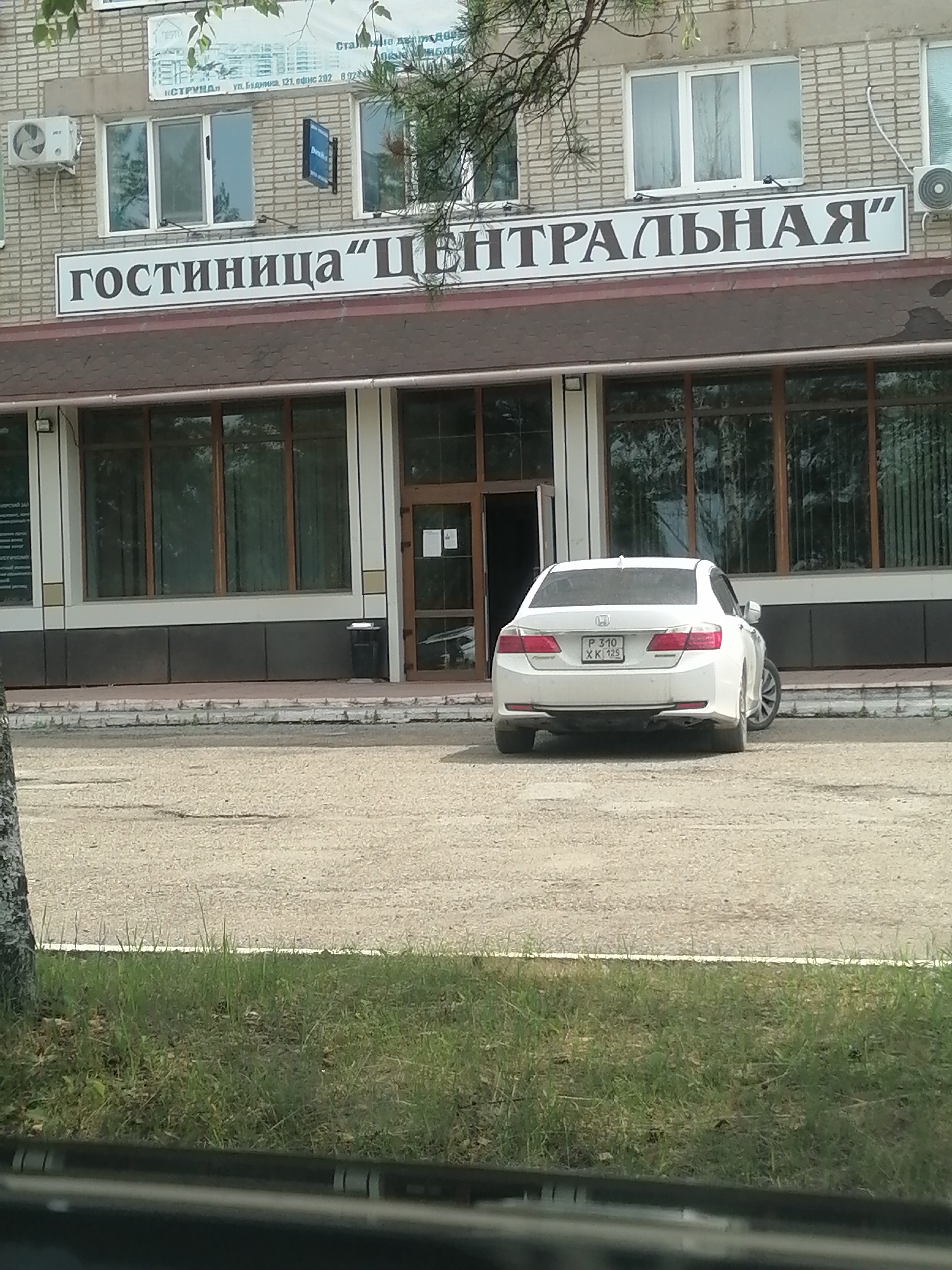 The image shows the facade of a brick building with large windows and a sign that reads "ГОСТИНИЦА "ЦЕНТРАЛЬНАЯ"" (Hotel "Centralnaya"). A white sedan is parked in front of the building. The building appears to be in a commercial or public area, with a paved area in front of it and some greenery. The license plate of the car is partially visible. The building is located in Lesozavodsk, Russia.