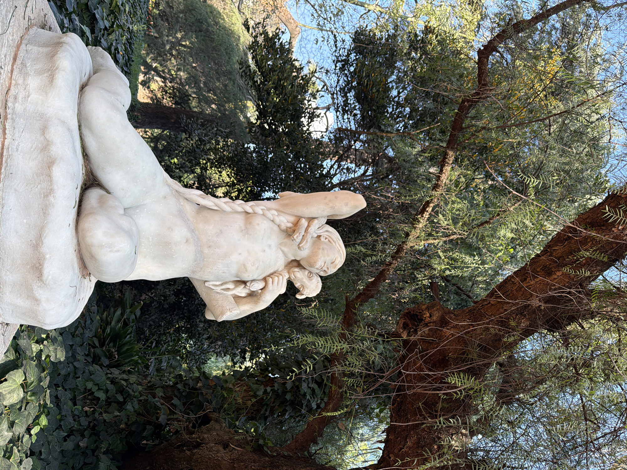 A white marble sculpture of a reclining figure with a sheep cradled in its arms is set on a stone pedestal. The figure has long braided hair, and one arm is raised towards the sky. The sculpture is situated in an outdoor park setting with lush green foliage and trees. The park appears to be the Rosedal de Palermo, located in Buenos Aires, Argentina. The sky above is a clear blue, indicating daytime. The sunlight casts gentle shadows, highlighting the texture of the sculpture and the surrounding vegetation.
