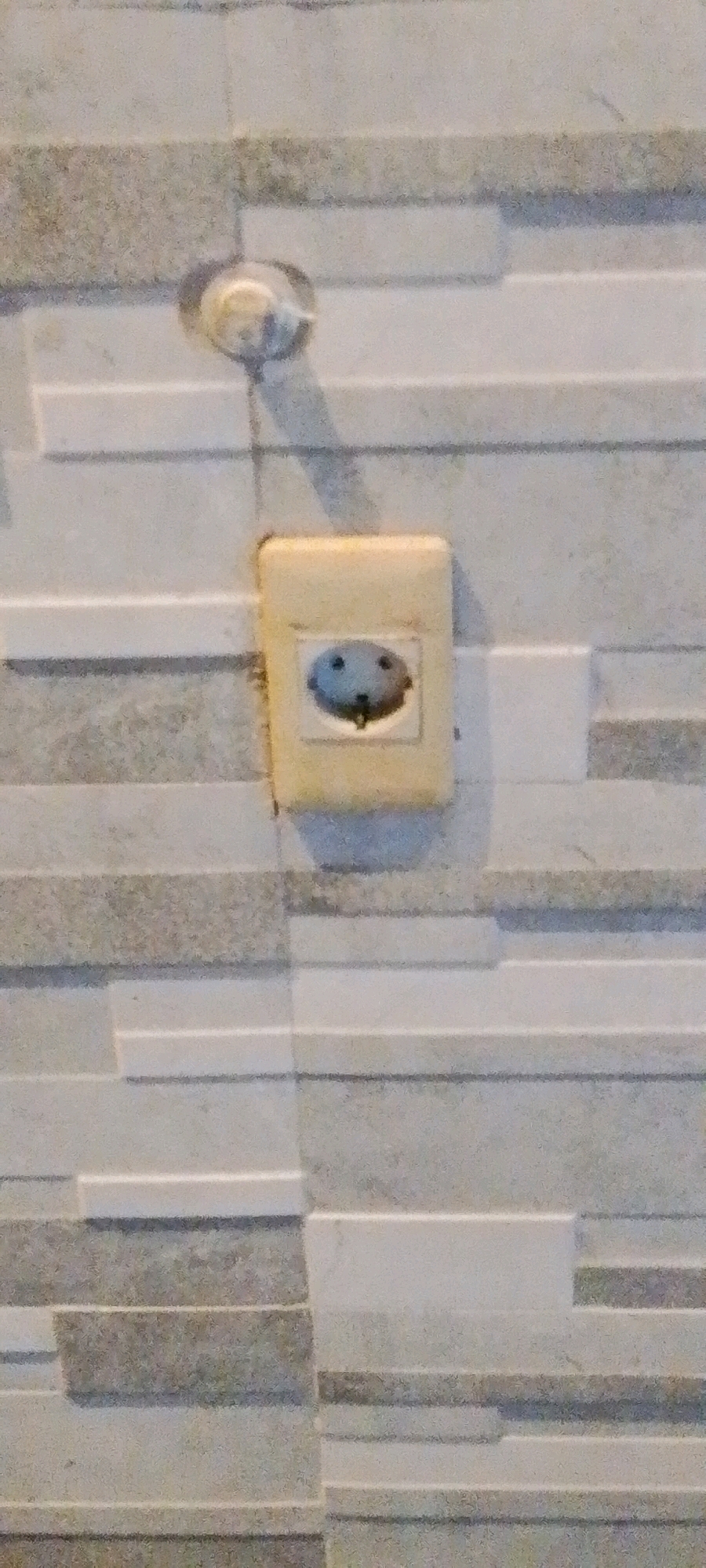 A close-up, eye-level shot shows a beige electrical outlet with a dark circular interior mounted on a wall covered with horizontal rectangular tiles. The tiles are arranged in a stacked pattern, alternating between light gray and darker gray textured stones. Above and slightly to the left of the outlet is a small, round, white fixture, possibly a light or a junction box cover. The lighting is dim, casting soft shadows on the wall. The image appears to be taken indoors. There are no people or animals visible in the frame.