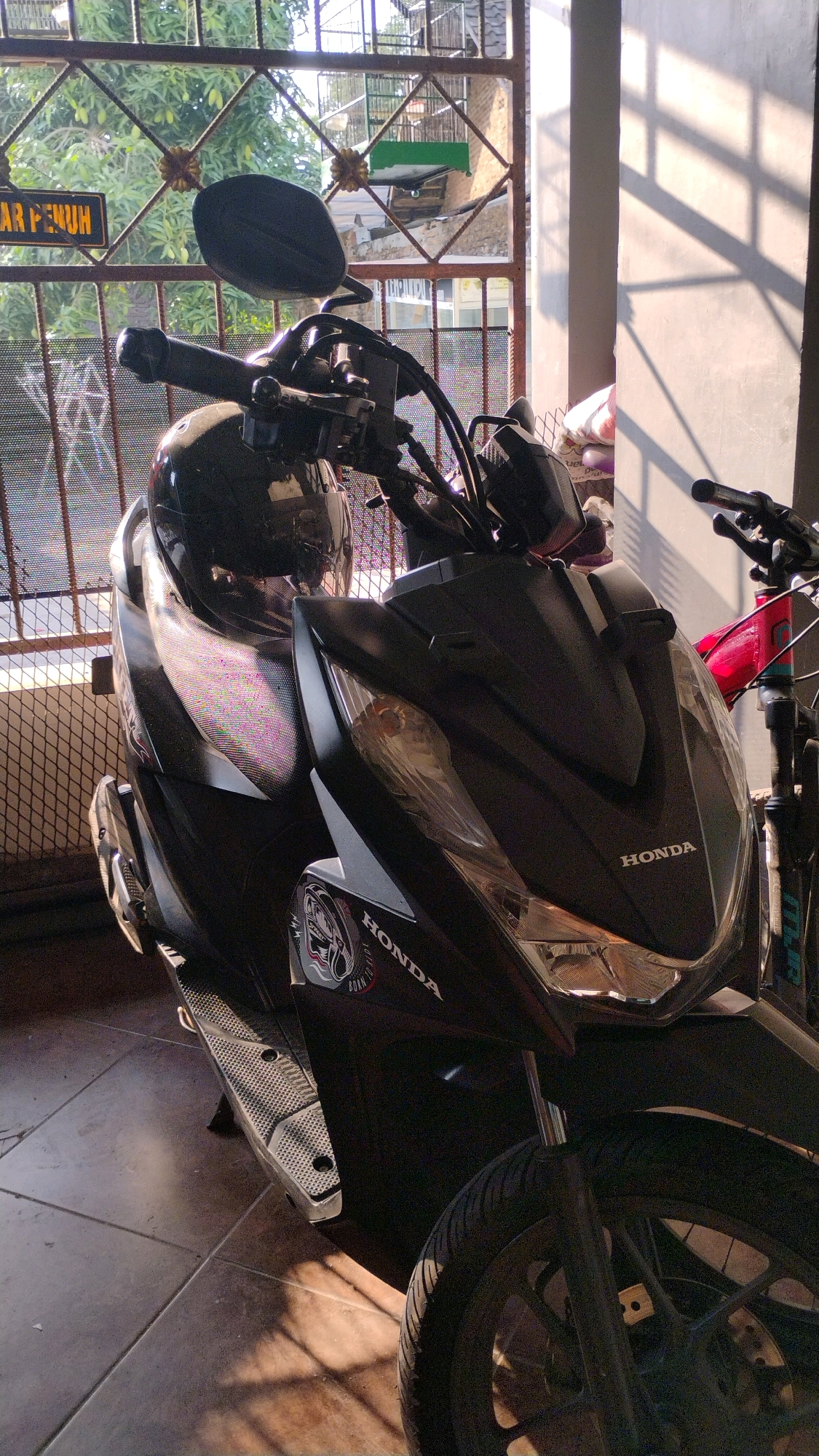 In Gumpang, Indonesia, a black Honda scooter is parked under a covered area, with a motorcycle helmet resting on the handlebars. The scooter is positioned in front of a fence that allows a view of trees and a brick building outside. Sunlight casts strong shadows across the concrete floor and a nearby wall. A bicycle with a red frame is partially visible to the right of the scooter. A sign with text, partially obscured, is visible on the fence. The scene appears to be a residential or semi-public outdoor space.