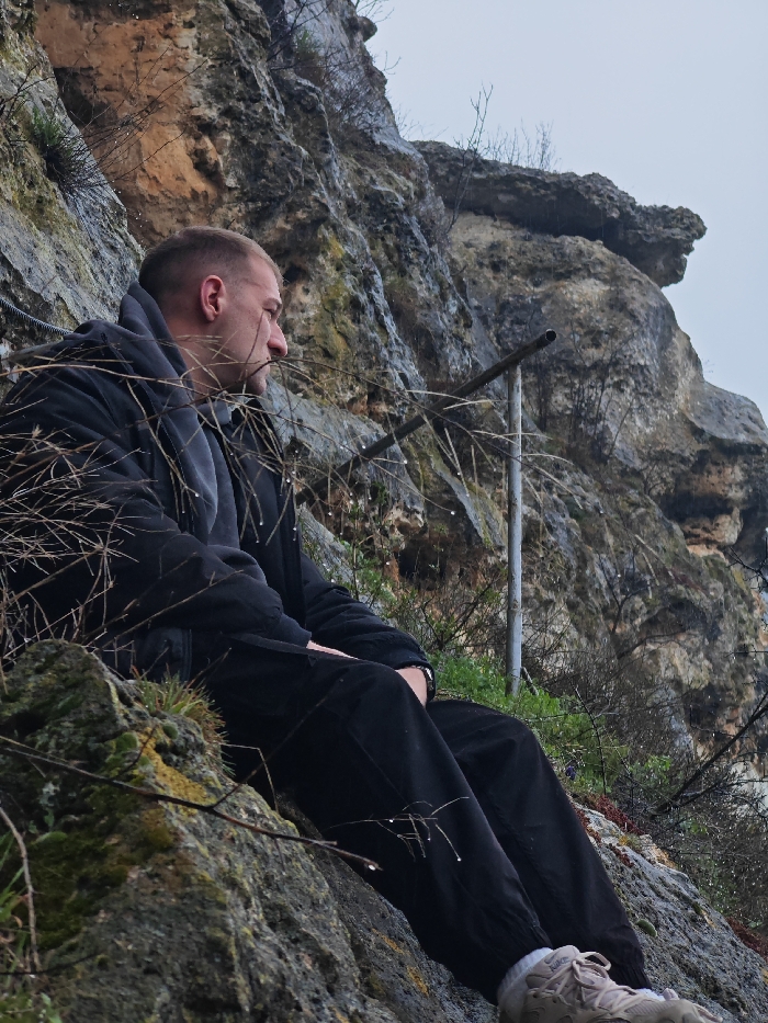A male individual with short, light-colored hair, wearing a dark jacket over a grey hooded garment, black pants, and light-colored athletic shoes with white socks, is seated on a rocky ledge. He is positioned facing right, with his head turned to the right, observing an unseen area beyond the frame. His right forearm is visible, sporting a dark wristband or watch. The setting is a rugged, multi-textured limestone cliff face with patches of moss and sparse, bare vegetation. Water droplets are visible on the branches and rocks, suggesting recent precipitation. In the upper left background, a steel safety cable is affixed horizontally to the rock. To the right of the subject, a grey metal post extends vertically from the ground upwards against the cliff. The sky is overcast. This scene is consistent with the natural rocky terrain found on the Shumen Plateau, Shumen, Bulgaria.