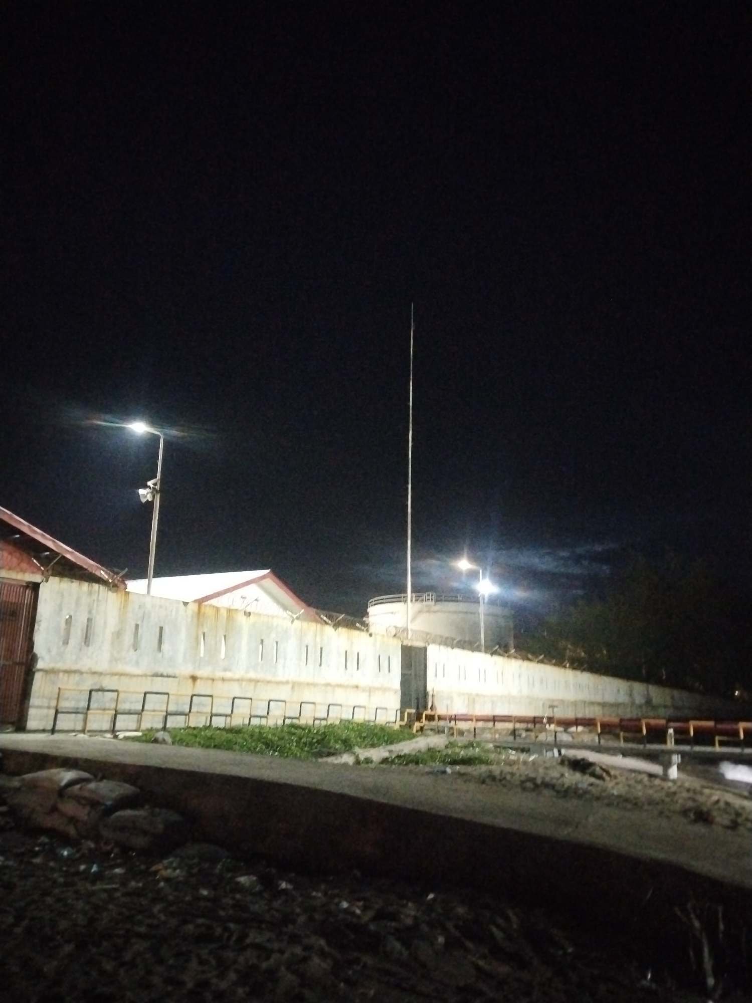 The image shows an industrial area at night in Lhokseumawe, Indonesia. A concrete wall with vertical rectangular indentations runs across the middle ground, with a red-roofed structure on the left and a large cylindrical tank on the right. Two streetlights illuminate the scene, one on the left casting a bright glow and another on the right near the tank, partially obscured by clouds. A tall, thin pole, possibly an antenna or flagpole, stands near the center. The foreground consists of dark, uneven ground with scattered debris and a raised concrete barrier. There are no people visible in the image.