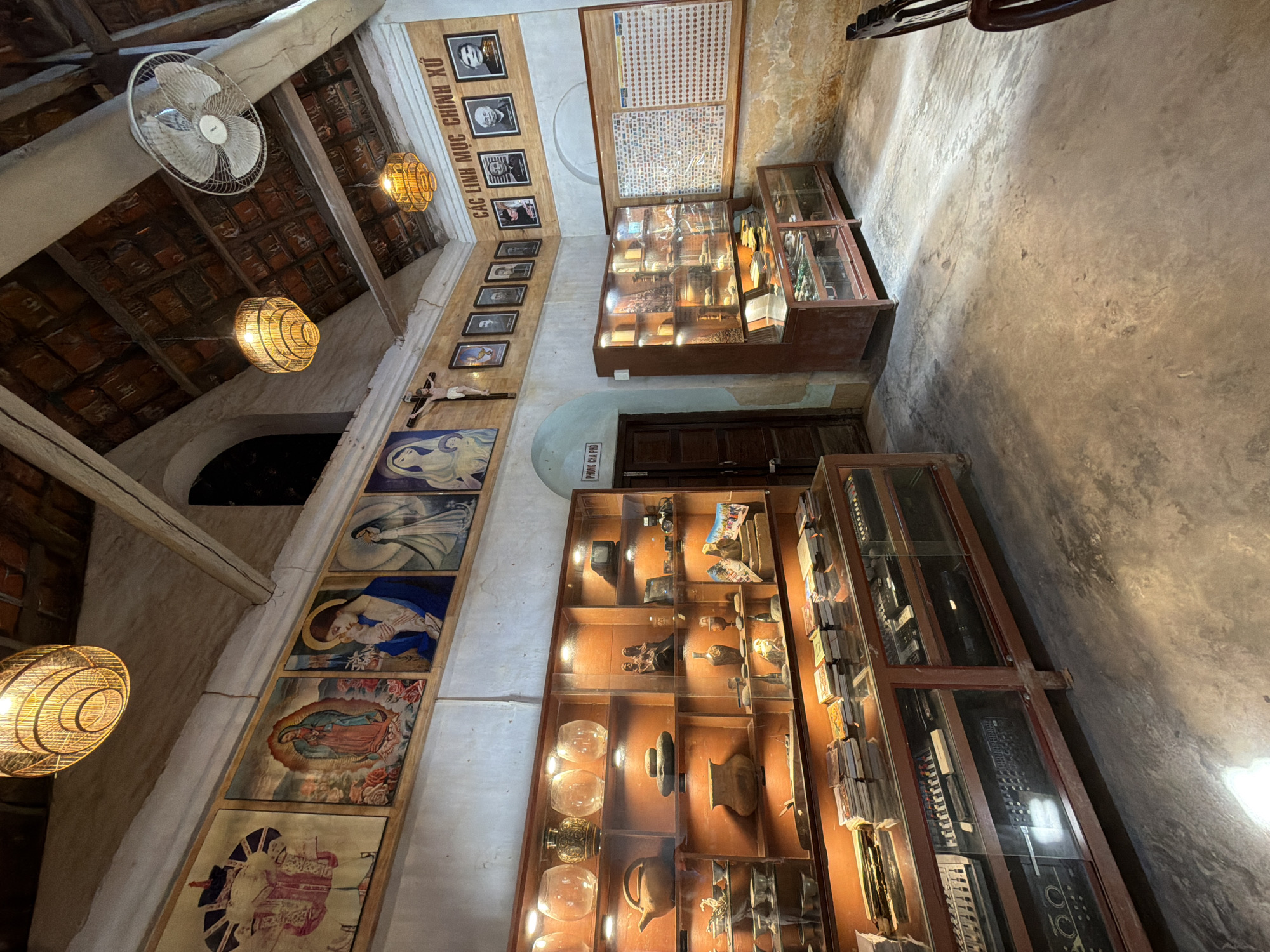 The image depicts the interior of a building that appears to be a museum or a religious exhibition space in Hội Tín, Vietnam. The room has a rustic aesthetic with exposed wooden beams and a tiled roof. Various religious paintings of what seem to be the Virgin Mary and other figures adorn the upper walls.
In the foreground, there are several display cases filled with artifacts. One large display case runs along the left wall, showcasing a collection of what appear to be vases, pottery, and possibly religious items. Below this, another long display case contains what looks like electronic equipment, possibly audio mixers or synthesizers, arranged neatly.
To the right, a taller display cabinet is filled with smaller objects, including what seem to be figurines and other decorative items. Adjacent to this cabinet, a large framed display board features a grid of numerous small photographs or stamps.
The space is illuminated by warm, artificial lighting from hanging lamps and spotlights within the display cases, casting shadows on the concrete floor. A wooden door is visible on the right side of the room, and a ceiling fan is mounted near the roof. The overall atmosphere is one of historical preservation and cultural display.