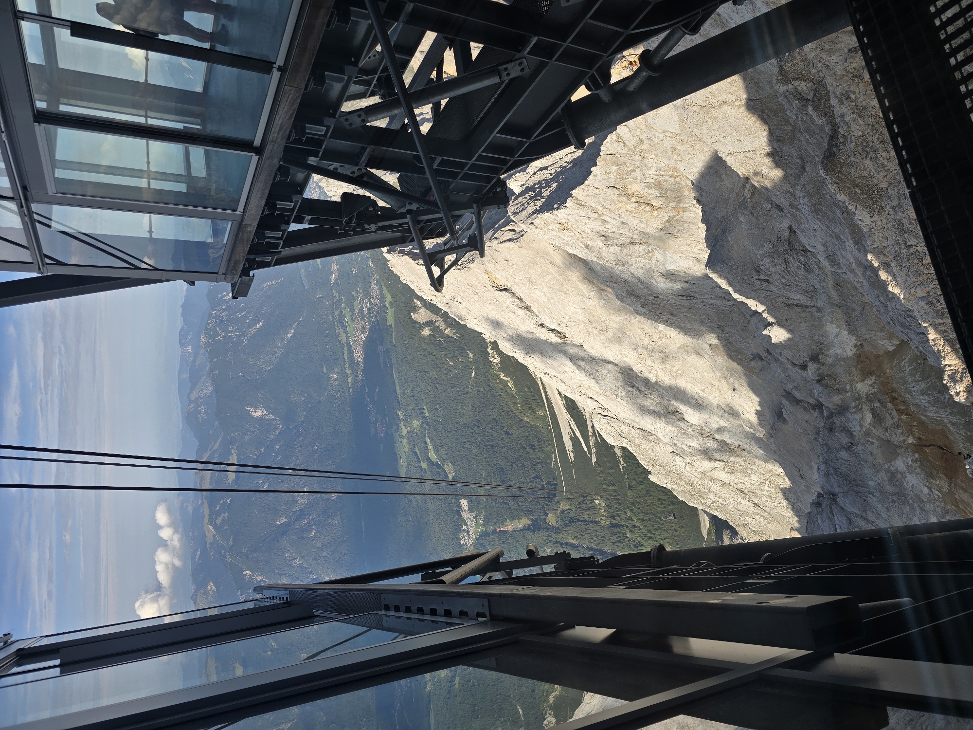 The image shows a view from a cable car station on a mountain, likely the Zugspitze near Garmisch-Partenkirchen, Germany. The foreground is dominated by the modern, glass-enclosed structure of the cable car station, with strong steel supports visible. Through the glass, the rugged, rocky surface of the mountain slopes down steeply. In the distance, a valley is visible with a dense green forest and scattered buildings, suggesting a lower elevation town or village. The sky is bright blue with some white clouds. The sunlight casts strong shadows on the mountainside, indicating it is likely daytime. One person is visible as a silhouette inside the cable car station, looking out at the view. The overall impression is one of a high-altitude tourist attraction offering dramatic views of the surrounding alpine landscape.
