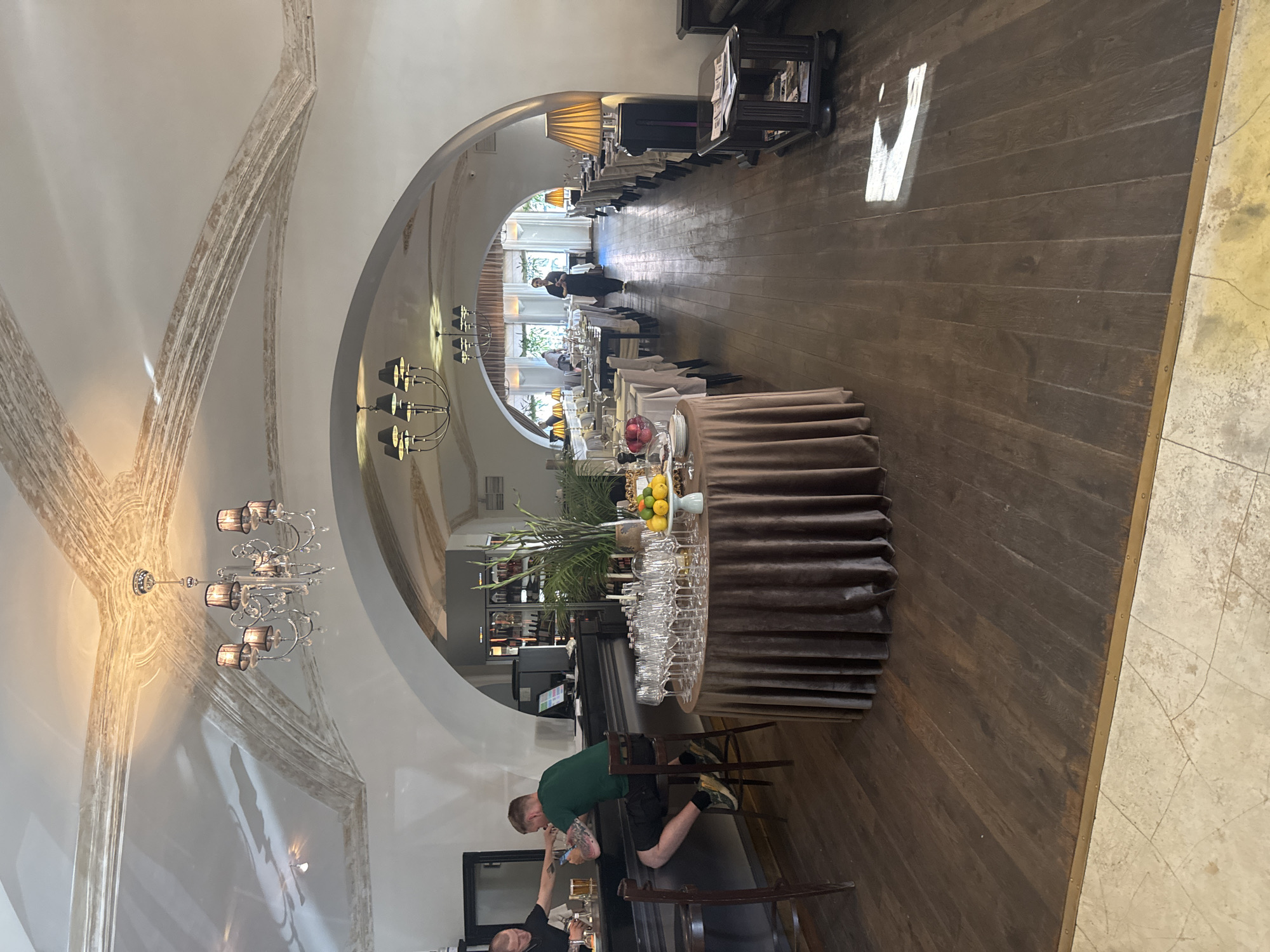 The image depicts the interior of a restaurant in Saint Petersburg, Russia, characterized by its arched ceilings adorned with decorative plasterwork and chandeliers. The dining area features numerous tables set with white tablecloths and glassware, arranged in rows. A bar area is visible on the left, with two individuals, possibly staff, interacting. One person is seated at the bar, while another stands behind it. A round table covered with a brown tablecloth, laden with glassware and a fruit display, is situated near the bar. Wooden flooring extends throughout the space. The overall ambiance suggests a formal dining establishment.