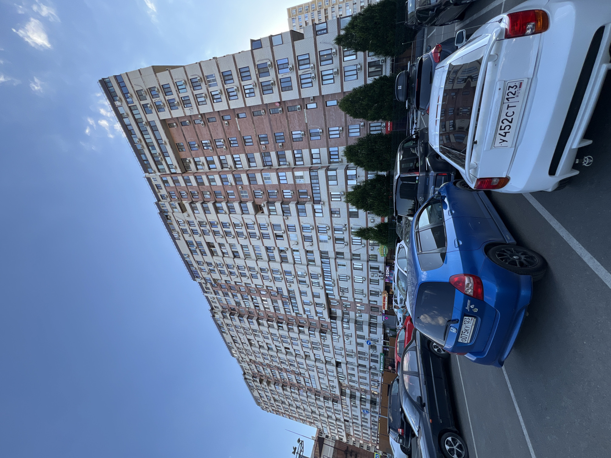 This is a wide-angle, eye-level, outdoor, daytime photograph of a large, multi-story apartment building in Krasnodar, Russia. The building, which appears to be of modern construction, has a long, angular facade with alternating sections of light-colored brick and red brick. Numerous windows and air conditioning units are visible across its many floors. In the foreground, a parking lot is occupied by several parked cars, including a blue hatchback, a white coupe, a black sedan, and a dark SUV. The sky above is clear blue with scattered wispy clouds. The scene captures a typical urban residential environment.