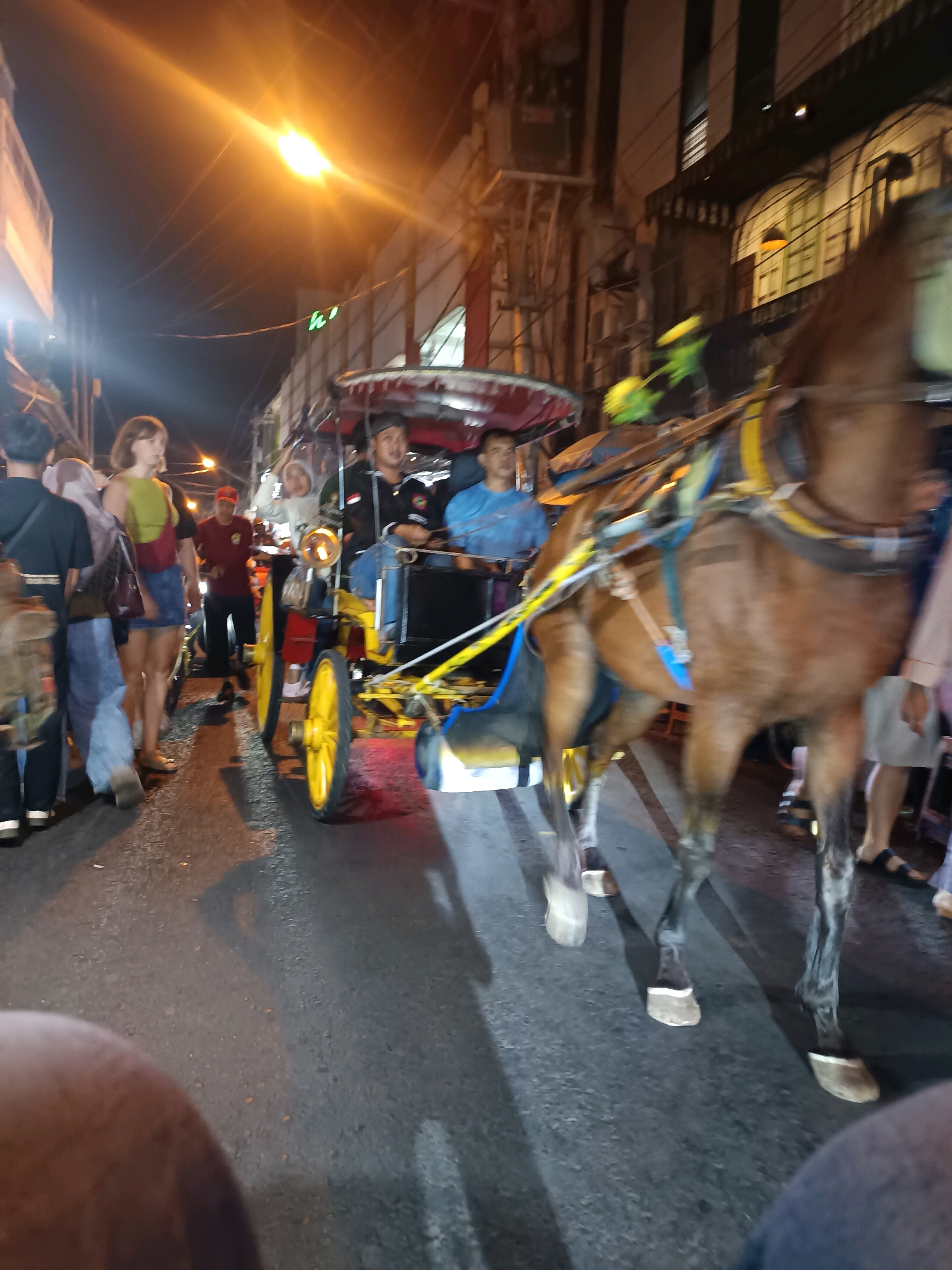 The image captures a night scene on a paved street in Yogyakarta, Indonesia, likely near a tourist area, with a horse-drawn carriage, known locally as a "andong," as the central subject. The andong, decorated with yellow wheels and colorful accents, is being pulled by a brown horse, which is positioned in the foreground and is partially blurred due to motion.
Several people are visible. Two men are seated in the andong, one appearing to be the driver, wearing a black jacket and a cap, and the other a passenger in a blue shirt. Several pedestrians are on the street, some walking and others standing. On the left, a woman in a green top and denim skirt is visible, with a dark bag slung over her shoulder. A man in a black shirt and another in a red shirt are also present. The street is illuminated by overhead lights, casting a warm glow on the scene. Buildings with visible windows and architectural details line the street, indicating an urban setting. The overall atmosphere suggests a lively evening activity, possibly a tourist attraction or a local event. The ground is wet in places, suggesting recent rain.