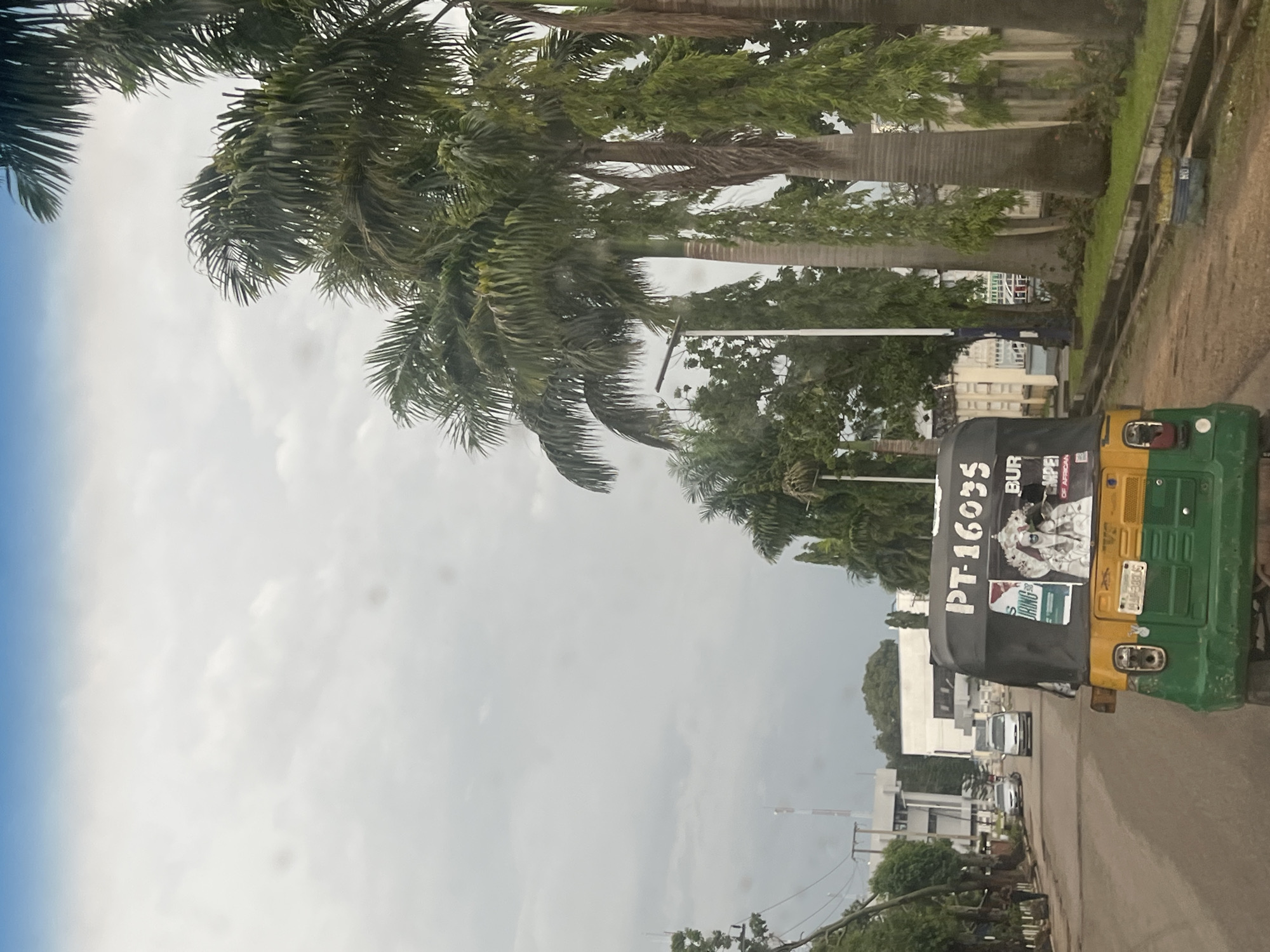 The image captures a street scene in Jos, Nigeria, featuring a three-wheeled motorized vehicle, commonly known as a "Keke Napep" or "Marwa." The Keke is predominantly yellow and green, with a black canopy displaying text. Visible text on the canopy includes "PT-16035," "BUR," and what appears to be a brand name or slogan partially obscured. Additionally, there are images on the canopy, including what seems to be a collage of people and potentially product advertisements. A license plate, "H/A 5835," is visible on the rear of the Keke.
The Keke is positioned on a road with a concrete drainage channel running alongside it. The road surface appears to be asphalt or a similar material. To the left of the road, there is a row of palm trees with lush green fronds, some of which are leaning over the road. Beyond the trees, there are buildings, including one with a white facade and multiple floors. The sky is overcast and appears to be a pale blue and white, suggesting a cloudy day. The overall lighting suggests it could be daytime, possibly late morning or afternoon, given the diffused light.
The scene depicts a typical urban or semi-urban environment in Nigeria. There are no people clearly visible in the immediate foreground, but the Keke suggests transportation activity. The presence of the Keke, palm trees, and architecture provides strong location cues for Jos, Nigeria. The weather appears to be mild, with no indication of rain, despite the overcast sky. The overall mood conveyed by the image is neutral, capturing a snapshot of daily life and transportation in the specified location.