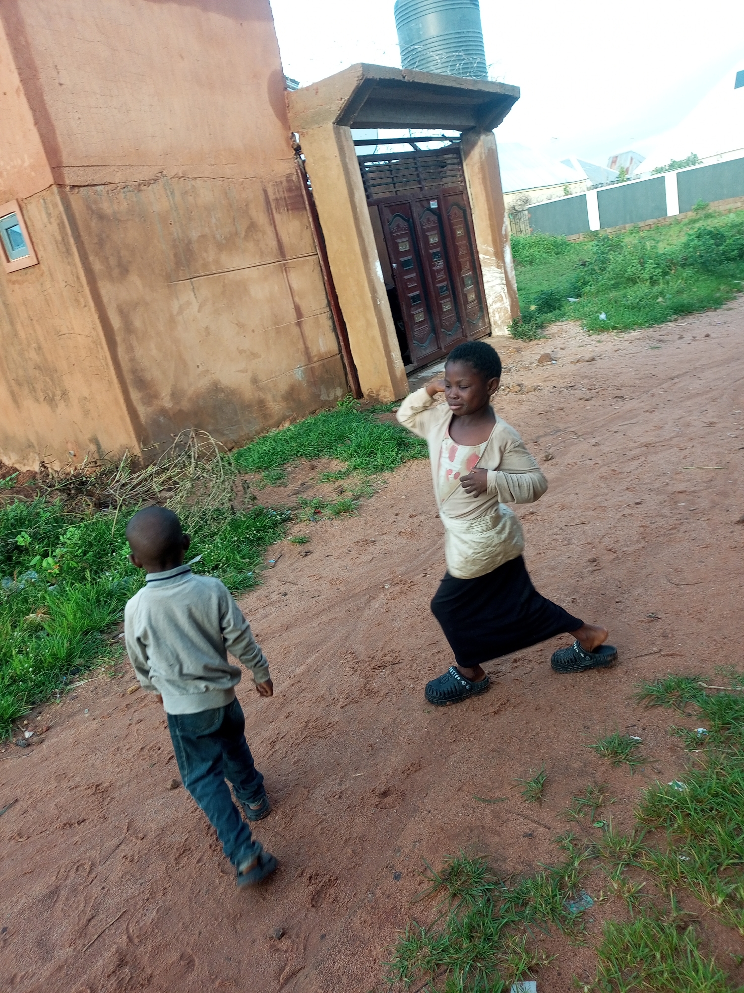 In Tempure, Nigeria, two children are standing on a dirt path beside a weathered, orange-colored building. A young girl, positioned to the right, is captured mid-movement, her right arm raised in a gesture that could be playful or part of a dance. She wears a light-colored long-sleeved shirt and a black skirt, with black clogs on her feet. To the left, a younger child, facing away from the camera, is dressed in a grey sweater and blue jeans, also wearing dark-colored footwear. The building features a small square window with a light blue frame. To the right of the building, a doorway with decorative brown doors is visible, topped by a concrete overhang. A large grey water tank rests on the roof of this section of the building. The ground is a mix of sandy dirt and sparse green grass. In the background, there is vegetation and what appears to be a wall or fence. The sky is overcast.