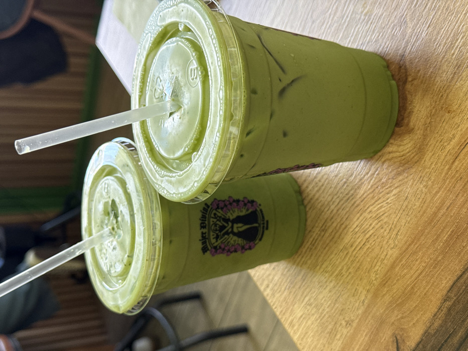 Two clear plastic cups containing iced light-green beverages are centered on a light brown wooden table with visible grain. Both cups are sealed with clear plastic dome lids and contain transparent plastic straws. The beverage in each cup contains ice cubes. The cup on the left features a label with the text "Mujer Divina" positioned above a dark silhouette figure framed by purple floral or wing-like designs, with smaller text "MATCHA & COFFEE" below. The background is out of focus, indicating an indoor setting, possibly a cafe or restaurant. The scene is located in Chula Vista, United States of America.