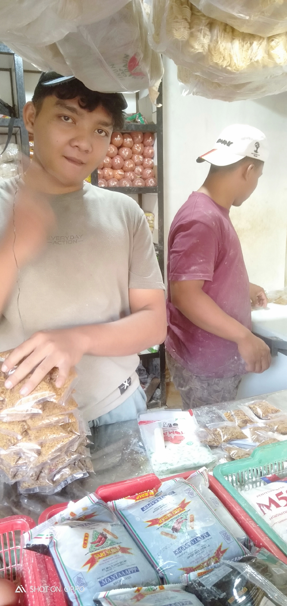 The image shows two men in what appears to be a food stall or small grocery store in South Jakarta, Indonesia. In the foreground, a young man wearing a dark t-shirt and a black baseball cap is holding up several small, clear plastic bags filled with a granular substance, possibly spices or dried food. He is looking towards the camera with a slight smile. Behind him, another man in a maroon t-shirt and a white baseball cap is seen from the back, appearing to be working or organizing items on a counter. The counter is covered with various packaged food items, including bags of what looks like instant noodles and other dried goods. Shelving units are visible in the background, stacked with more products, including what seems to be an abundance of round, reddish-brown items in clear packaging. The overall environment suggests a busy, small-scale retail setting where food products are stored and sold. The presence of the brand "Sari Sedap" on some of the packaging is noted. The lighting suggests an indoor environment, possibly with overhead fluorescent lights. The surfaces appear dusty, indicating a potentially active workspace.