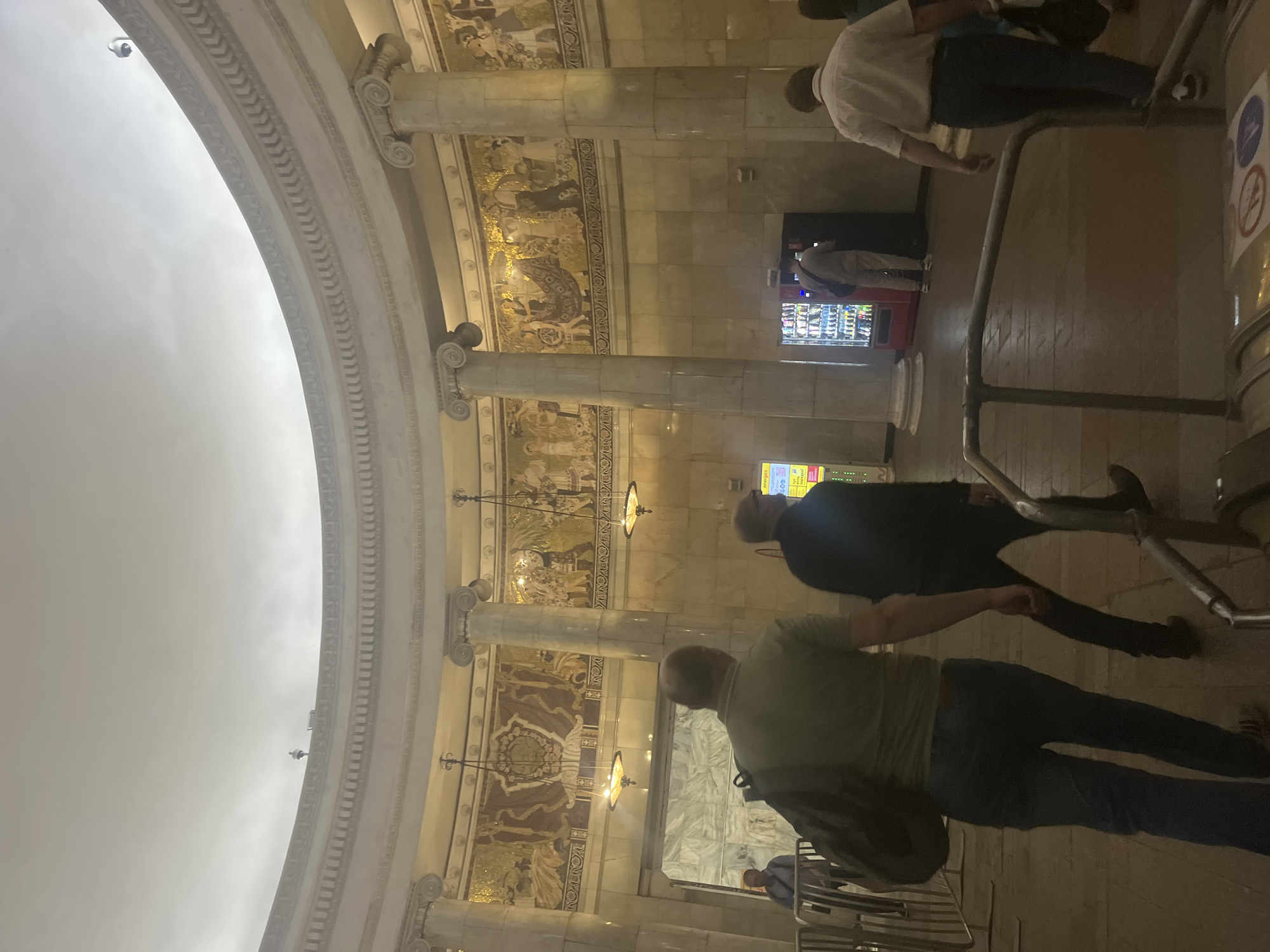 The image captures the interior of a grand public space, strongly resembling a Moscow Metro station, characterized by its elaborate architecture and decorative elements. The scene is oriented vertically, showing a high, curving arched ceiling adorned with intricate classical moldings, including a security camera mounted high up.
The walls below the arch are richly decorated with detailed mosaic or fresco panels, depicting various scenes featuring human figures, animals, and possibly agricultural or mythological motifs. These artworks are framed by robust, fluted columns with ornate capitals, characteristic of classical design. Ornate chandeliers hang from the ceiling, casting a warm light.
Several people are visible, engaged in typical transit activities. In the foreground, a person wearing a light green shirt, blue jeans, and a backpack walks towards the right, away from the viewer. Another person in a dark jacket and pants walks alongside them. Further into the scene, a person in a white shirt and dark pants moves in the same direction. Near a marble-paneled wall, a red vending machine with illuminated products is visible, and a person in grey attire stands near it.
To the far right, metal turnstiles or railings suggest an entry or exit point. A yellow sign partially visible on the right displays Cyrillic text, including "ВНИМАНИЕ" (Attention) and "тарифы" (tariffs), indicating informational content. Circular signs, including a clear "No Smoking" symbol, are mounted near the turnstiles. The overall impression is of a well-lit, bustling, and historically rich transit environment during active hours.