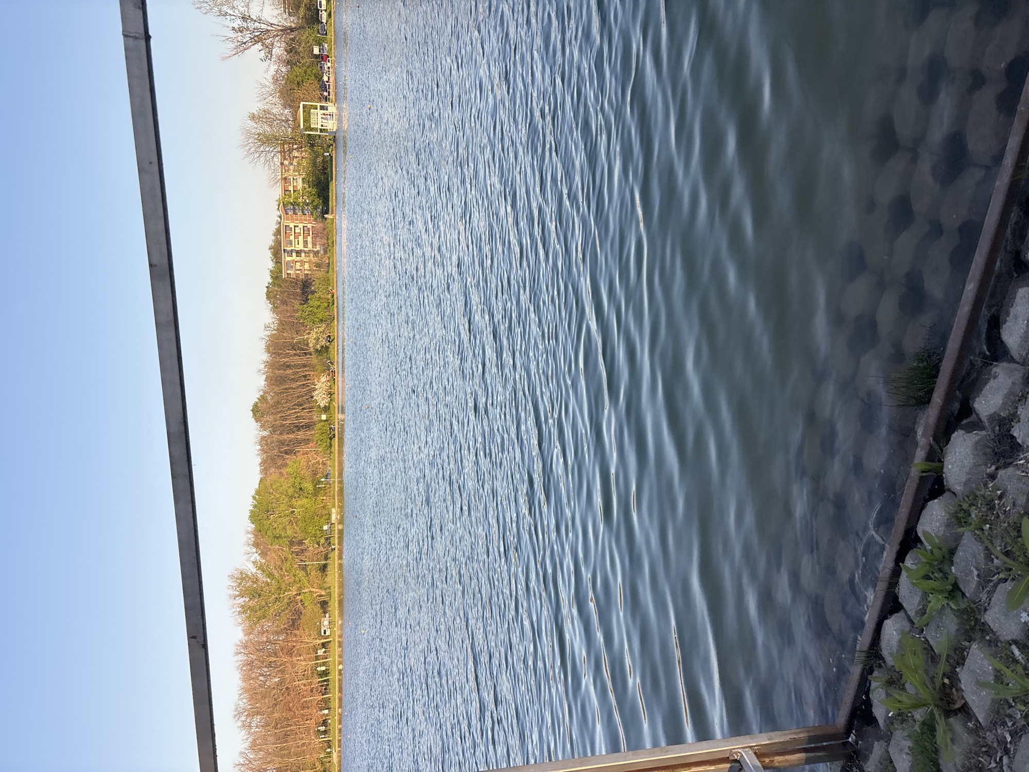 The image shows a body of water, likely a lake, with a rippled surface. Along the right edge, there's a structure made of concrete blocks or stones, with some vegetation growing on them. In the background, across the water, there's a line of trees, some buildings, and what appears to be a park structure. The sky is visible along the left edge of the frame. The image could be taken at the Rowing Canal in Plovdiv, Bulgaria, given the presence of a large body of water and surrounding park-like environment.