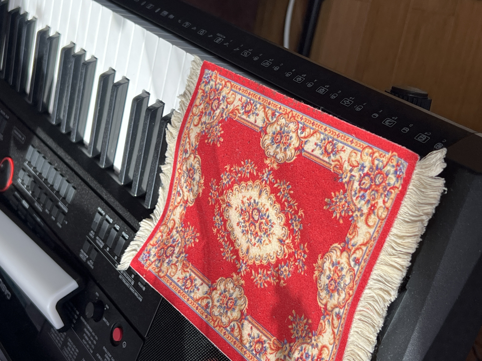 A close-up shot shows a Casio electric piano with white and black keys. A small, red decorative rug with a floral pattern and fringed edges lies on the piano. The piano's control panel is partially visible with various buttons and labels such as "Volume," "Rhythm/Song Controller," "Intro," and "Variation/B." The keyboard is positioned at an angle, giving a perspective view of the keys receding into the background. The background is out of focus, showing a hint of a wooden surface or wall. The location is not evident from this image.
