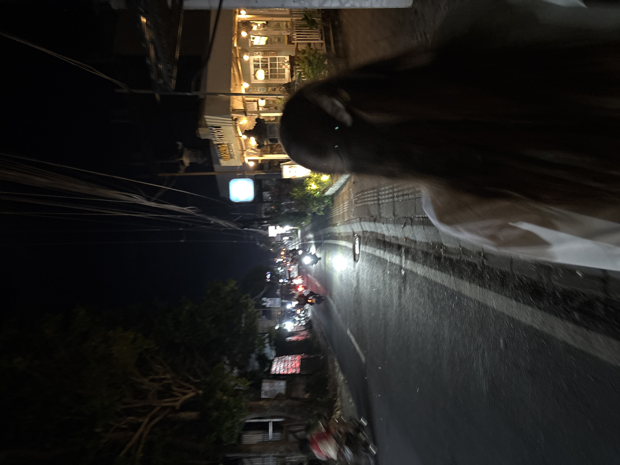 A nighttime scene captured along a street in Ungasan, Indonesia. A person is walking along the side of the road. The street is asphalt, marked with a white line dividing the lanes. Several motorcycles are traveling down the road. Buildings line the street; some are illuminated. Wires are visible overhead. The focus is soft in the foreground, with greater clarity toward the center of the frame.