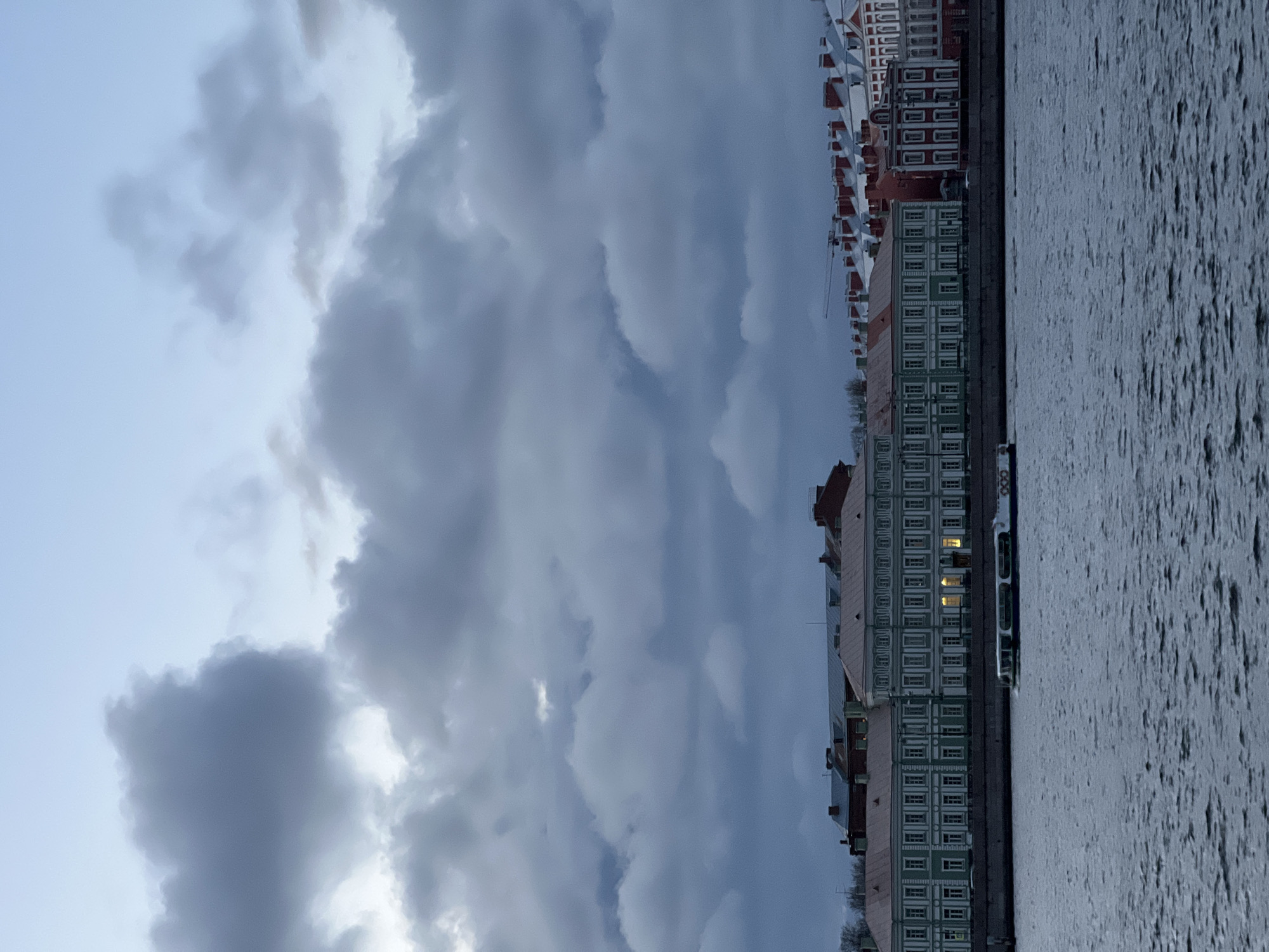 The image depicts a cityscape in Saint Petersburg, Russia, likely taken from across the Neva River. The sky is overcast with heavy, textured clouds covering most of the frame. Along the riverbank, a row of buildings with varying colors and architectural styles stands. The buildings have multiple floors and numerous windows, with some windows lit. The Neva River fills the right side of the frame with visible water surface texture.