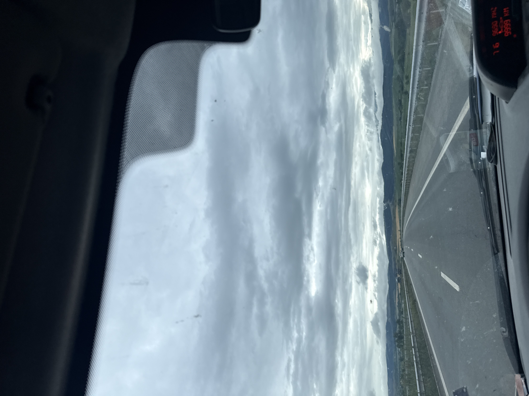The image captures a view from inside a vehicle traveling on a highway. The sky is overcast with dense, grey clouds. The road ahead is paved with asphalt and has white lane markings. Guardrails line the right side of the highway, separating it from a grassy embankment and fields beyond. In the distance, rolling hills and mountains are visible, some with patches of green vegetation and others appearing bare or rocky. A few other vehicles are seen on the highway in the distance, and the overall impression is of a travel scene under a cloudy sky. There is a digital display on the vehicle's dashboard showing "9999 KM". The location is stated to be in the city of Delyan, Bulgaria.