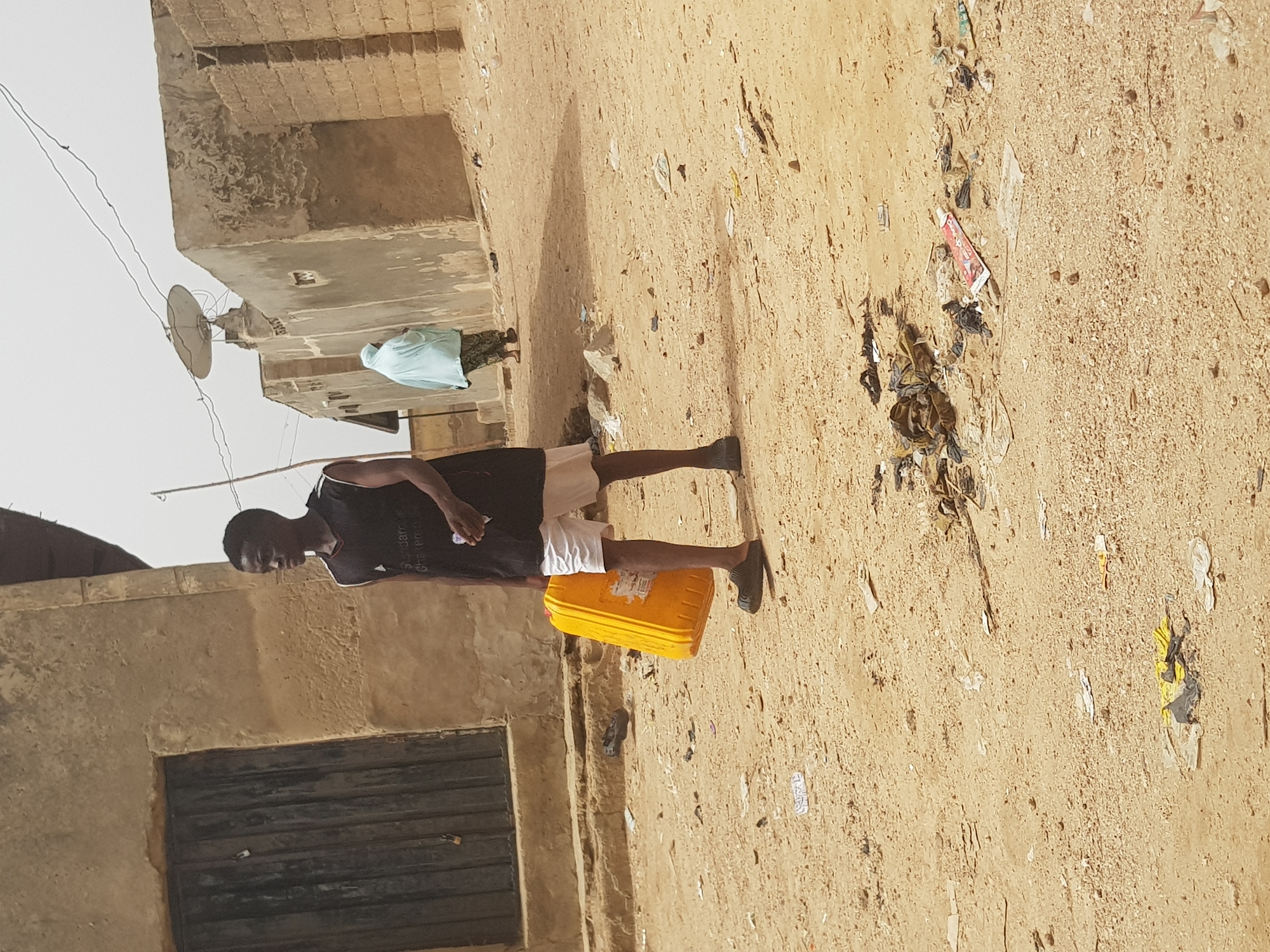 An eye-level, long shot shows a man walking along a dirt road in Dutsinma B, Nigeria. The man, dressed in a dark T-shirt and light shorts, carries a yellow container. The ground is dirt covered with litter. Buildings are to the left, and another person is further down the road in the distance.
