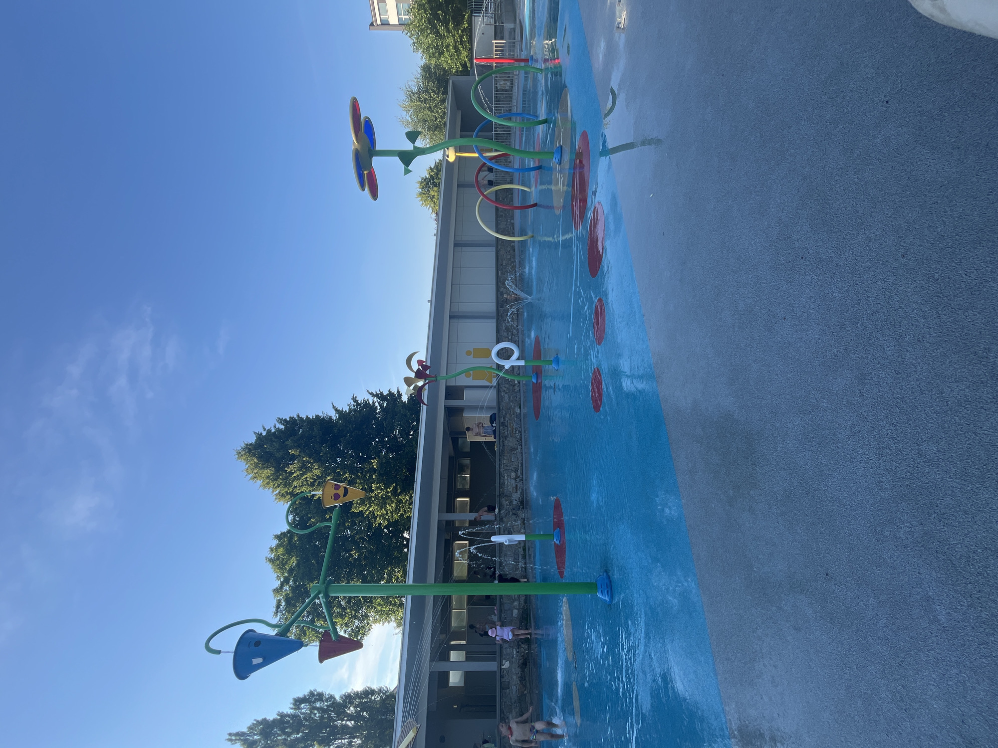 A water playground is shown on a sunny day with a clear blue sky and scattered clouds. The playground features several water features, including sprayers, buckets, and arches, all set within a shallow pool with bright blue water. The surrounding area is paved with concrete, with some sections colored red. In the background, a building with a row of windows and a stone facade is visible. Several people are present, including children playing in the water and adults watching. The overall impression is one of a recreational space on a pleasant day.