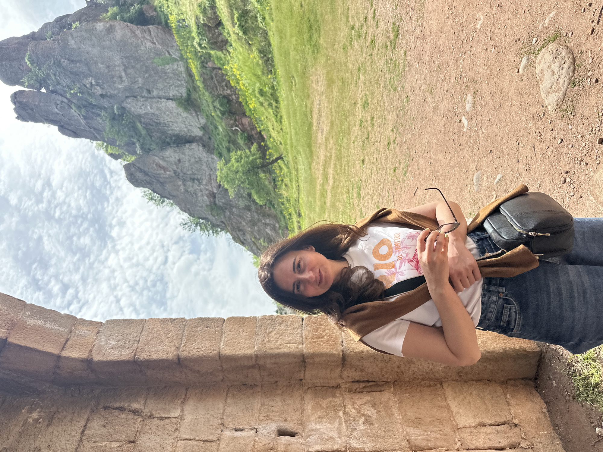 A person stands in Belogradchik, Bulgaria. The individual is a woman, facing the camera with a slight smile, wearing a white t-shirt, blue jeans, and a brown jacket draped over her shoulders. She holds a pair of sunglasses in her hands. A black bag hangs on her right hip. The woman leans against a stone wall of tan or light brown color. The wall is constructed of rectangular stone blocks. In the background is a landscape of greenery and a large, rocky formation. The sky is visible with white clouds. A dirt path or area is seen on the ground.