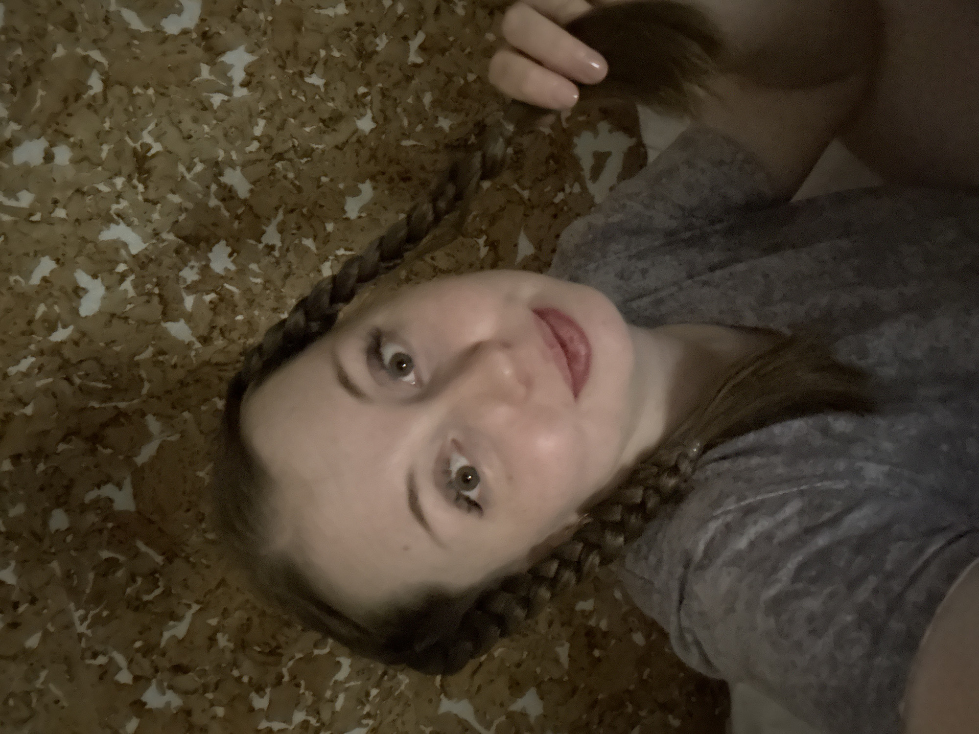 A young woman with light skin and light brown hair is depicted in a close-up, tilted shot, looking upwards towards the camera. She appears to be lying down, possibly on her side or back. Her hair is styled in two braids that frame her face, and her right hand (viewer's left) is visible in the upper part of the frame, gently holding a section of her unbraided hair. She is wearing a casual grey t-shirt.
Her facial expression is playful and cheerful, characterized by a slight smile and the tip of her tongue peeking out. Her brown eyes are wide open and directly engaging with the viewer. The background consists of a textured surface, possibly cork or a similar material, featuring an irregular pattern of light-colored flecks against a darker brown base, suggesting an indoor setting. The lighting is soft and somewhat dim, contributing to an intimate and relaxed atmosphere. There is no visible text in the image.