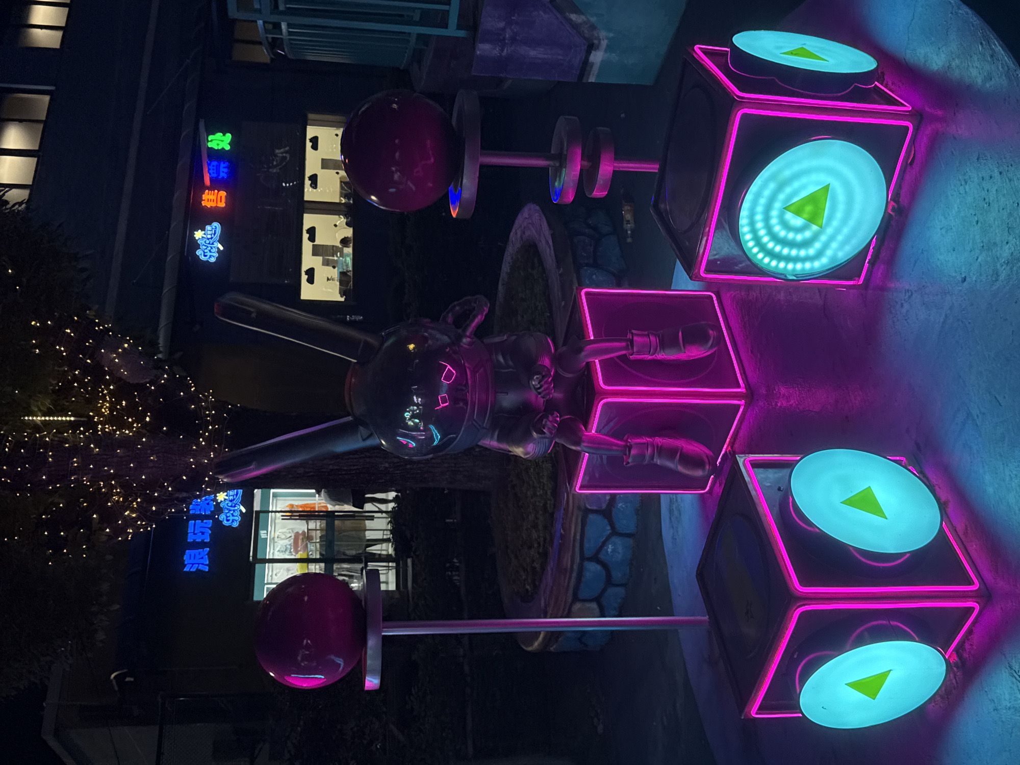 The image depicts a nighttime scene in Yiwu, China, likely in an outdoor public space or commercial area. In the foreground, three illuminated cubes are arranged, each with pink neon outlining and circular teal lights displaying green triangular shapes. A metallic sculpture of a seated rabbit-like character with glowing blue eyes sits atop one of these cubes. To the left and right of the rabbit sculpture are tall stands topped with reflective red spheres. In the background, a building with signage in Chinese characters is visible, illuminated by various neon lights. The building appears to be a storefront or commercial establishment. A tree decorated with strings of small lights is also present, casting a glow on the scene. The ground surface is a dark, reflective material. The overall atmosphere is characterized by vibrant neon lighting and a futuristic or playful aesthetic.