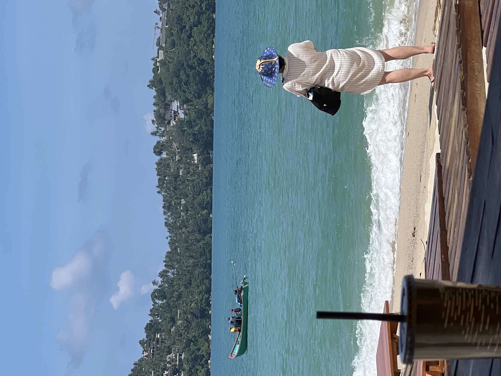 An eye-level, medium shot captures a woman standing on a wooden platform facing a turquoise ocean. She is wearing a wide-brimmed blue and yellow patterned hat, a light-colored striped dress, and black sandals. A black bag hangs from her left shoulder. She is positioned with her back to the camera, looking out at the water. In the middle ground, a small green boat with four people aboard is moving across the sea. The boat has a long pole extending from its stern. The background features a lush green hillside covered in dense trees and scattered buildings. The sky is a bright blue with scattered white clouds. In the foreground, a portion of a brown cup with a black straw is visible on the left side of the frame. Wooden planks of a deck or walkway are in the immediate foreground and extend to the right. Gentle waves are washing onto the sandy beach.