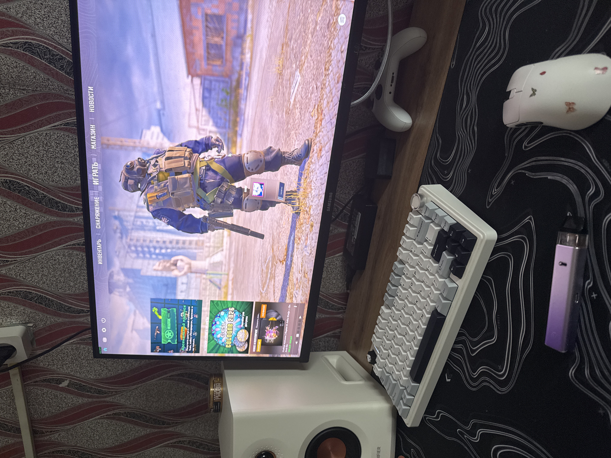 The image shows a gaming setup on a wooden surface with a black and white topographic patterned mouse pad. On the pad are a white gaming mouse decorated with butterflies, a white mechanical keyboard, and a purple vape device. A white gaming controller lies on the wood surface near the monitor. In the background, a monitor displays a first-person shooter video game featuring a character in tactical gear in an urban setting. On the bottom of the screen, there are game menu options in Russian. A white speaker sits in front of the monitor. A small item labelled "PHOBIA" is also present. The location is not specified in the image, and there is no indication of Perm, Russia.
