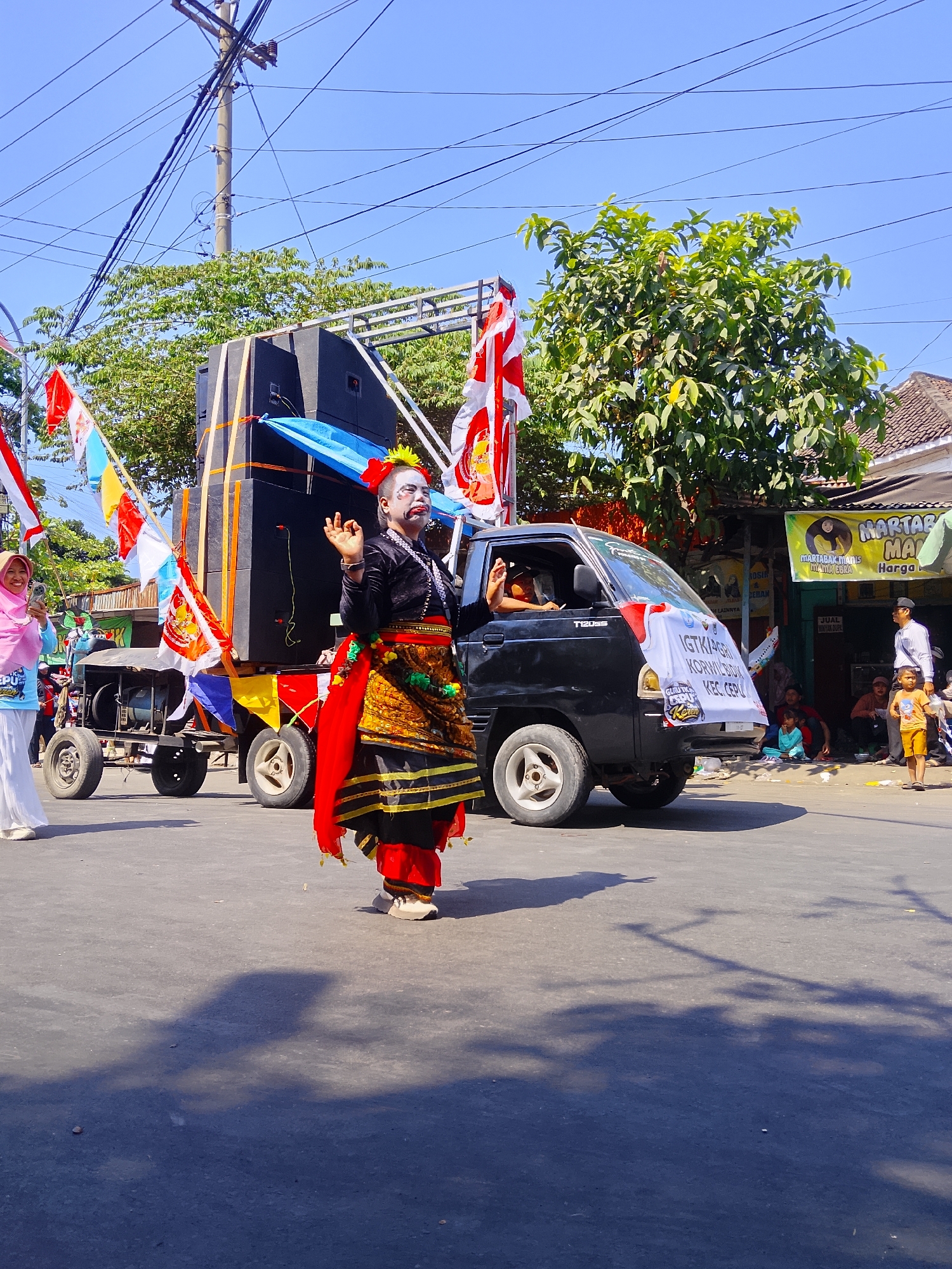 In the city of Balun, Indonesia, a street parade is underway on a sunny day. The foreground is dominated by the shadow of a tree cast across the asphalt road. To the right, a black pickup truck with a banner that reads "IGTKI-PGRI KORWILDA" is prominently featured. A person in elaborate traditional attire, with face paint and a red, gold, and black costume, is performing or posing in front of the truck. Behind them, a trailer carries a large sound system and is decorated with Indonesian flags and colorful pennants. To the left, a woman in a pink hijab and white clothing is seen holding a phone, possibly taking pictures of the parade. Spectators are gathered along the side of the road, with some sitting and others standing, observing the procession. The environment includes a utility pole with numerous wires overhead, trees providing shade, and buildings lining the street, one of which has a banner advertising "MARTABAK MANIS." The sky is clear and blue.