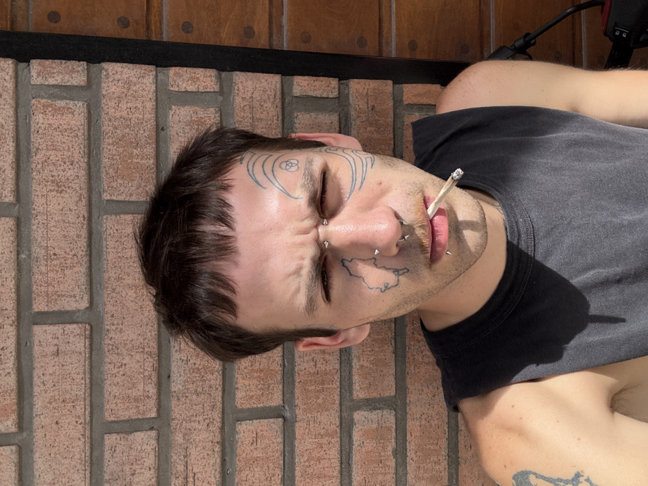 A person lies on brickwork, facing upward. The individual has short, dark hair and several facial piercings and tattoos. One tattoo is on the forehead and appears to be a geometric design, another is a depiction of a land mass on the cheek. The person holds a lit cigarette in their mouth. They wear a dark gray t-shirt, revealing their upper torso. A wooden structure is partially visible in the background, along with a black cable.